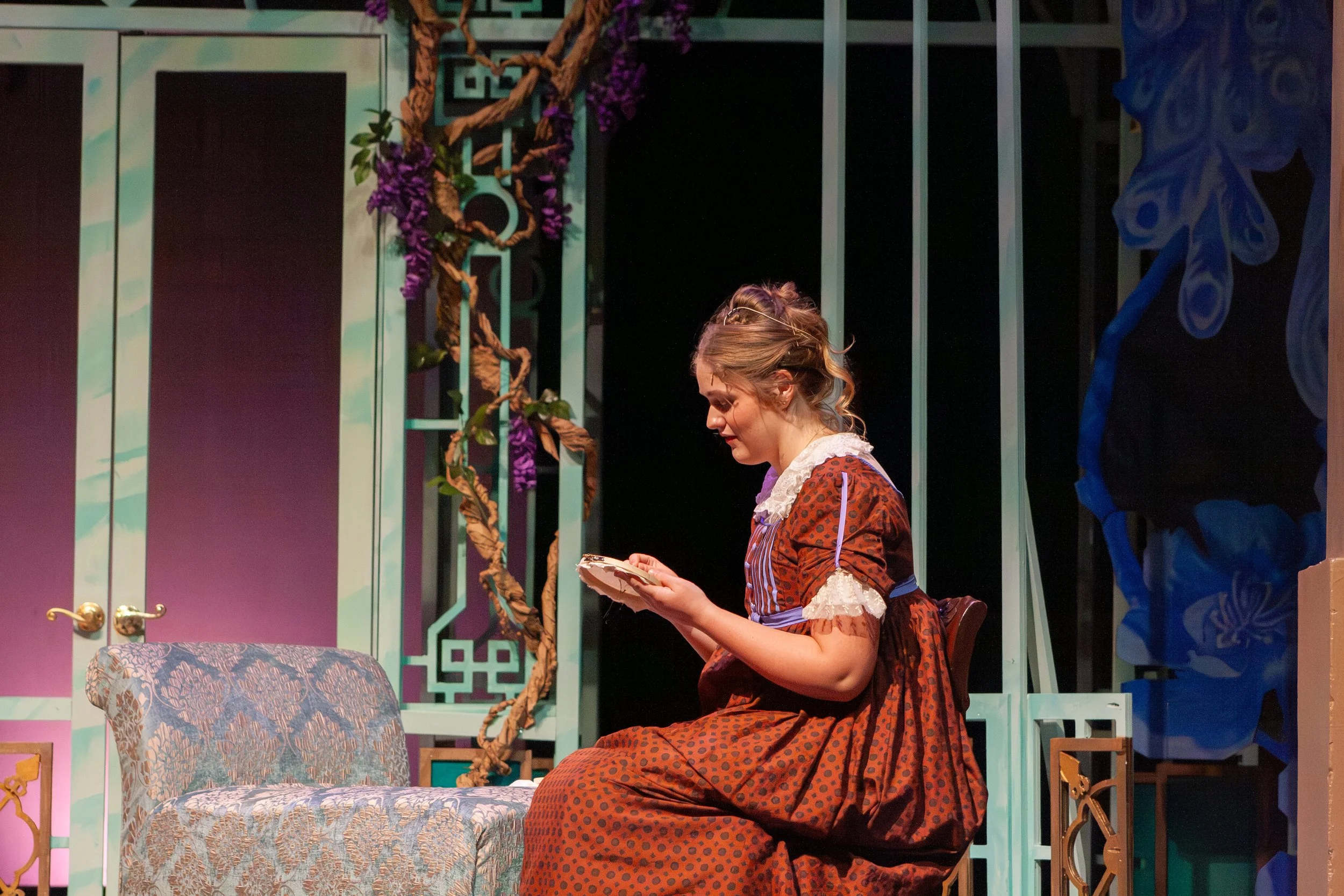 A woman dressed in a period costume, sitting on a chair on a theater stage, reading a book.