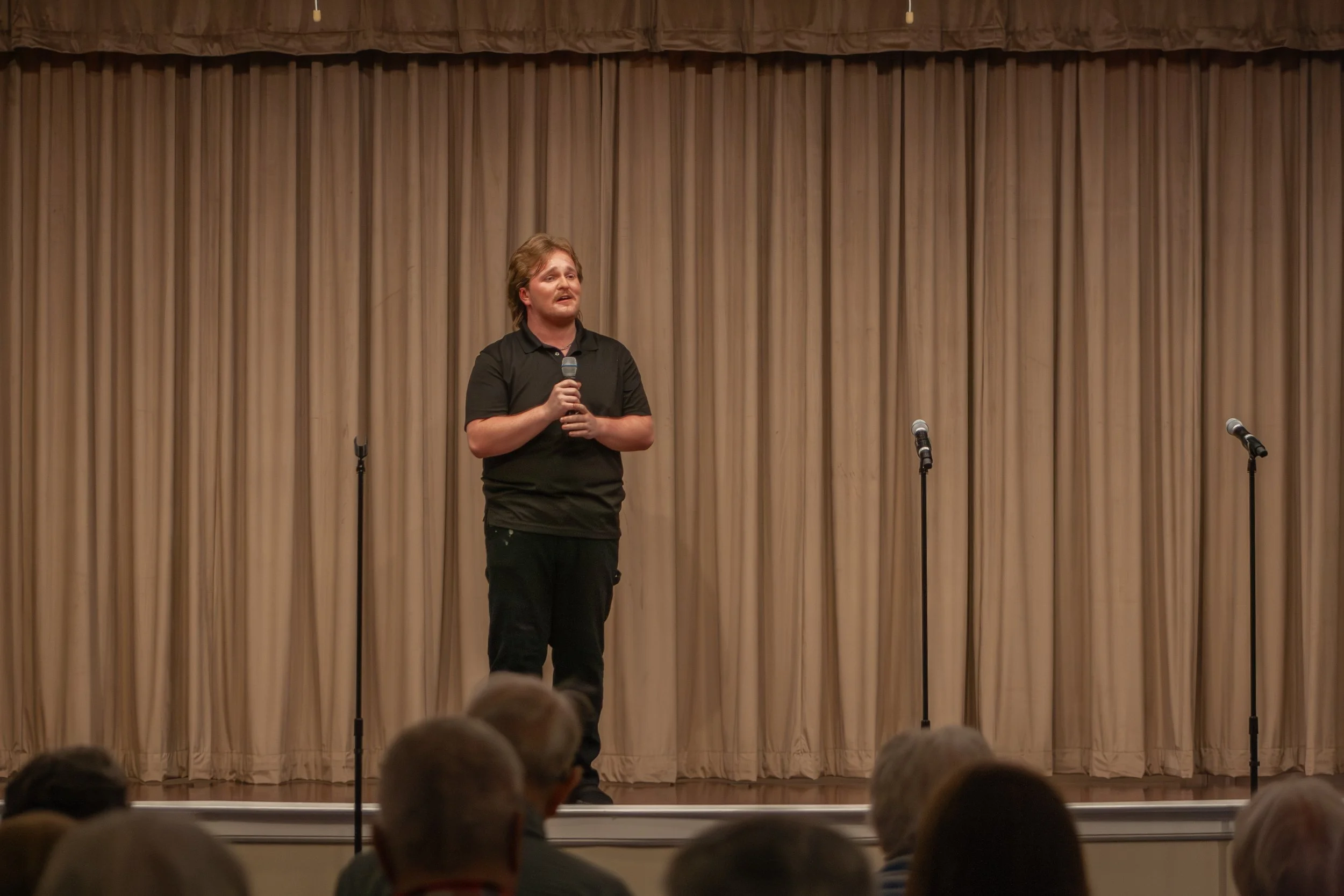 A man with light brown hair and a beard, wearing a black shirt, holding a microphone, speaking on stage in front of a beige curtain, with microphones on stands nearby, in front of an audience.