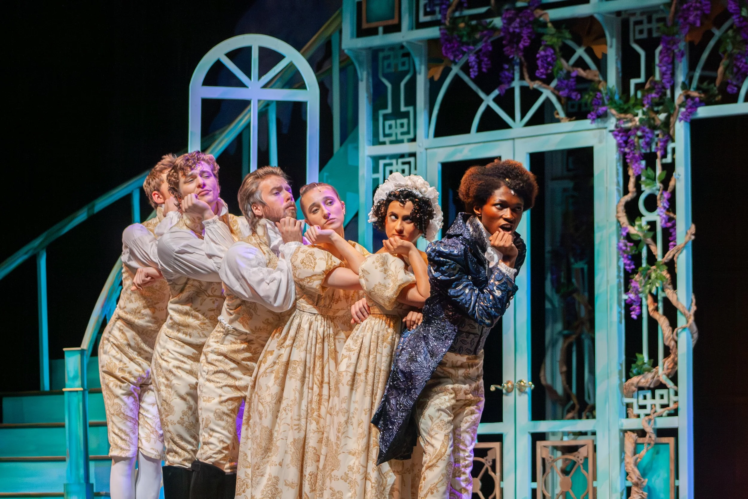 A theatrical stage scene featuring six performers dressed in historical costumes, standing in a line with their chins resting on their hands, with a blue and purple decorated set background.