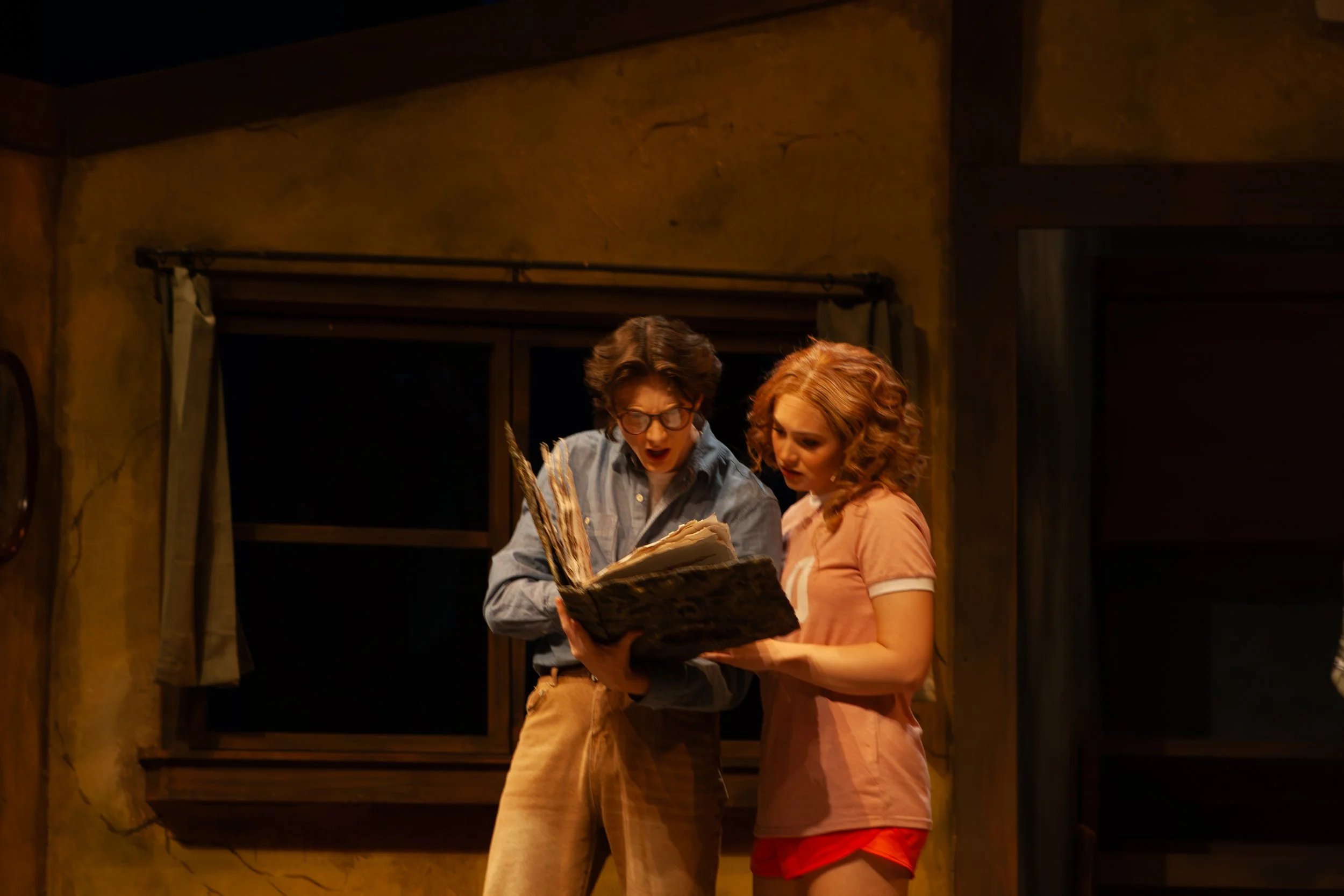 Two young people, a boy and a girl, are looking at a large book together in a rustic room with yellowish walls and a window.