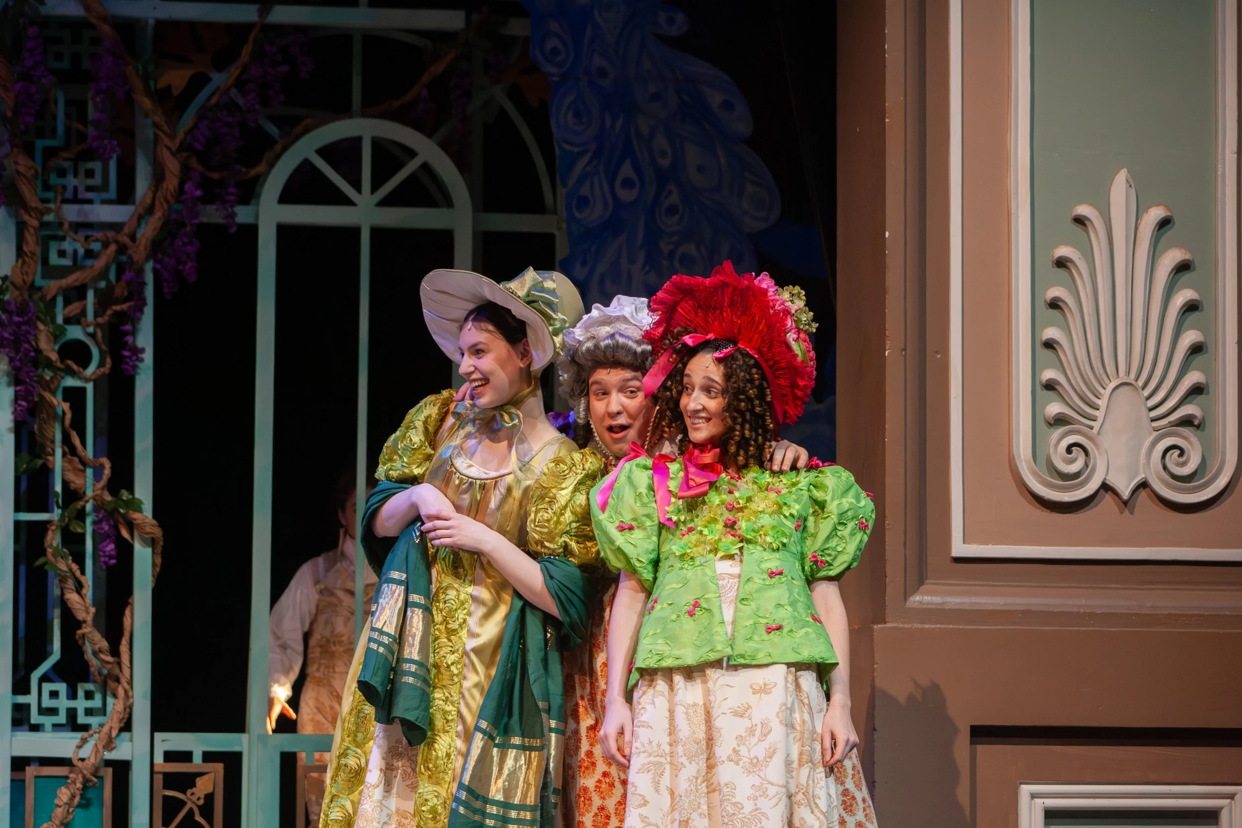 Three women in colorful period costumes standing close together on stage. They are smiling and appear to be in a theatrical performance, with a decorative set background behind them.