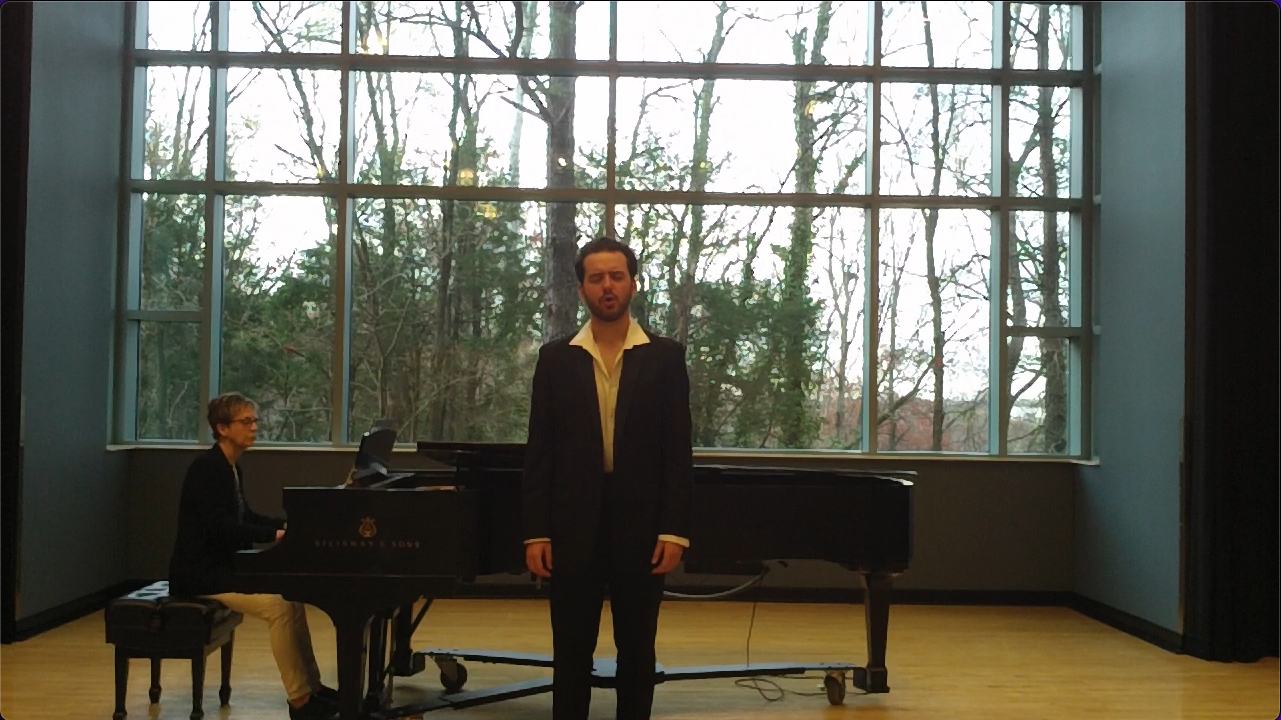 A man in a black suit singing in front of a large window with trees outside, while a woman plays the piano nearby.