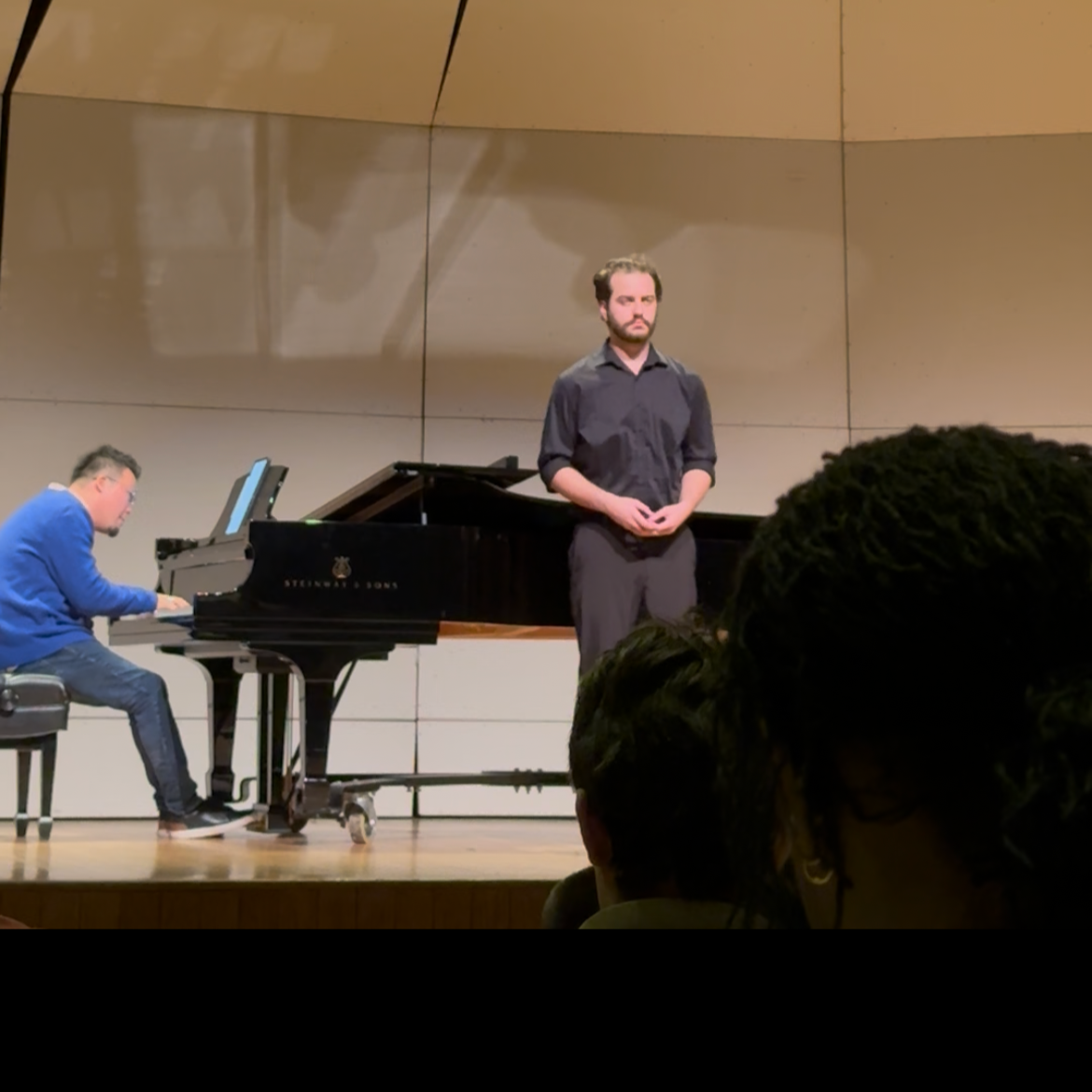 A man stands on stage in front of a piano, while another man plays at the piano. The man standing has a serious expression and is dressed in black, the pianist is wearing a blue sweater.