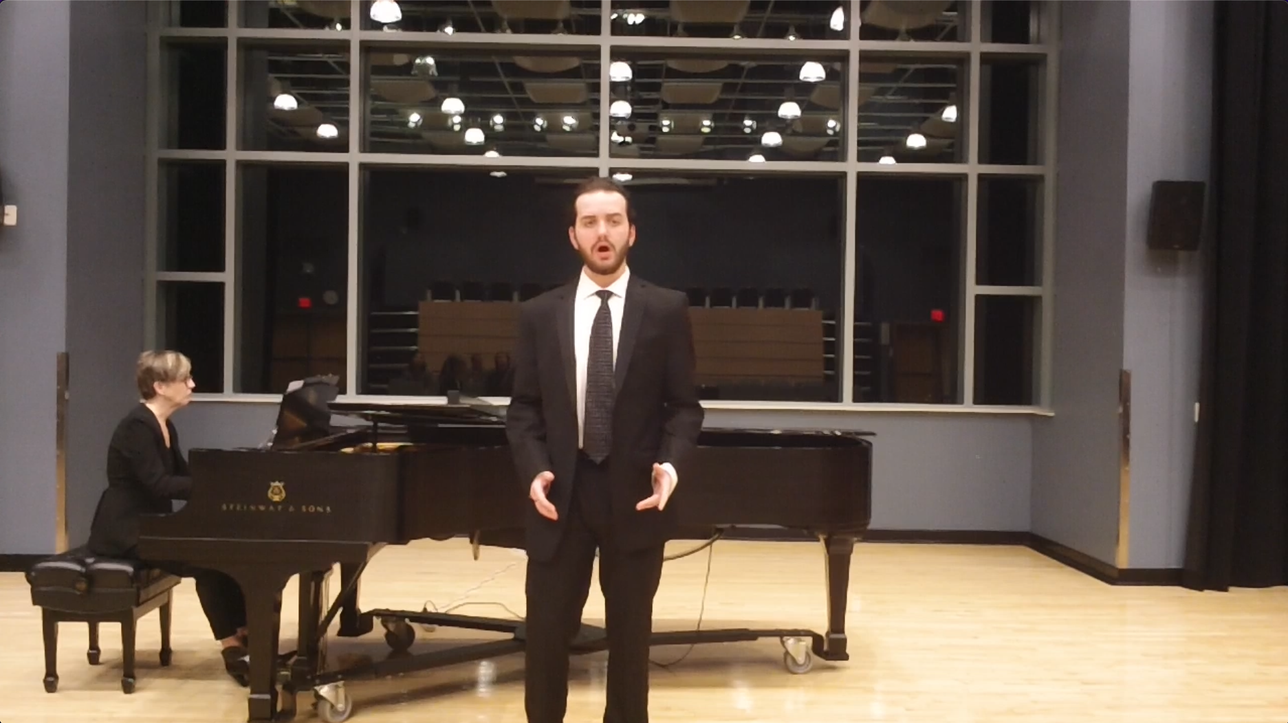 A male performer in a suit singing in front of a pianist playing a grand piano during a performance or rehearsal, in a room with large windows and a visible audience.