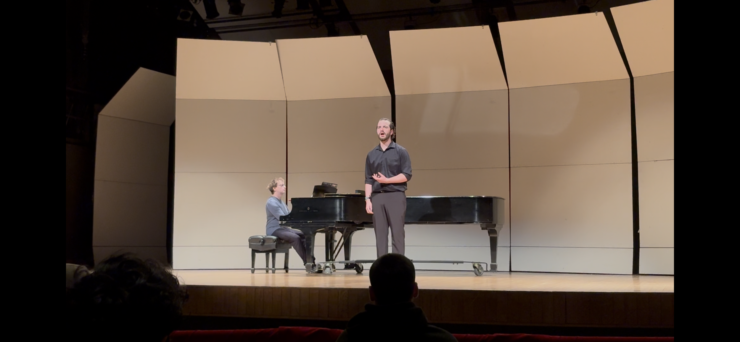 A man and a woman performing a piano recital on stage. The woman is sitting at a grand piano, while the man is standing and singing or speaking in front of the piano. They are in a concert hall with a light-colored, geometric wall backdrop.