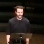 A man with a beard and dark hair standing behind a music stand or lectern, speaking or preparing to speak.