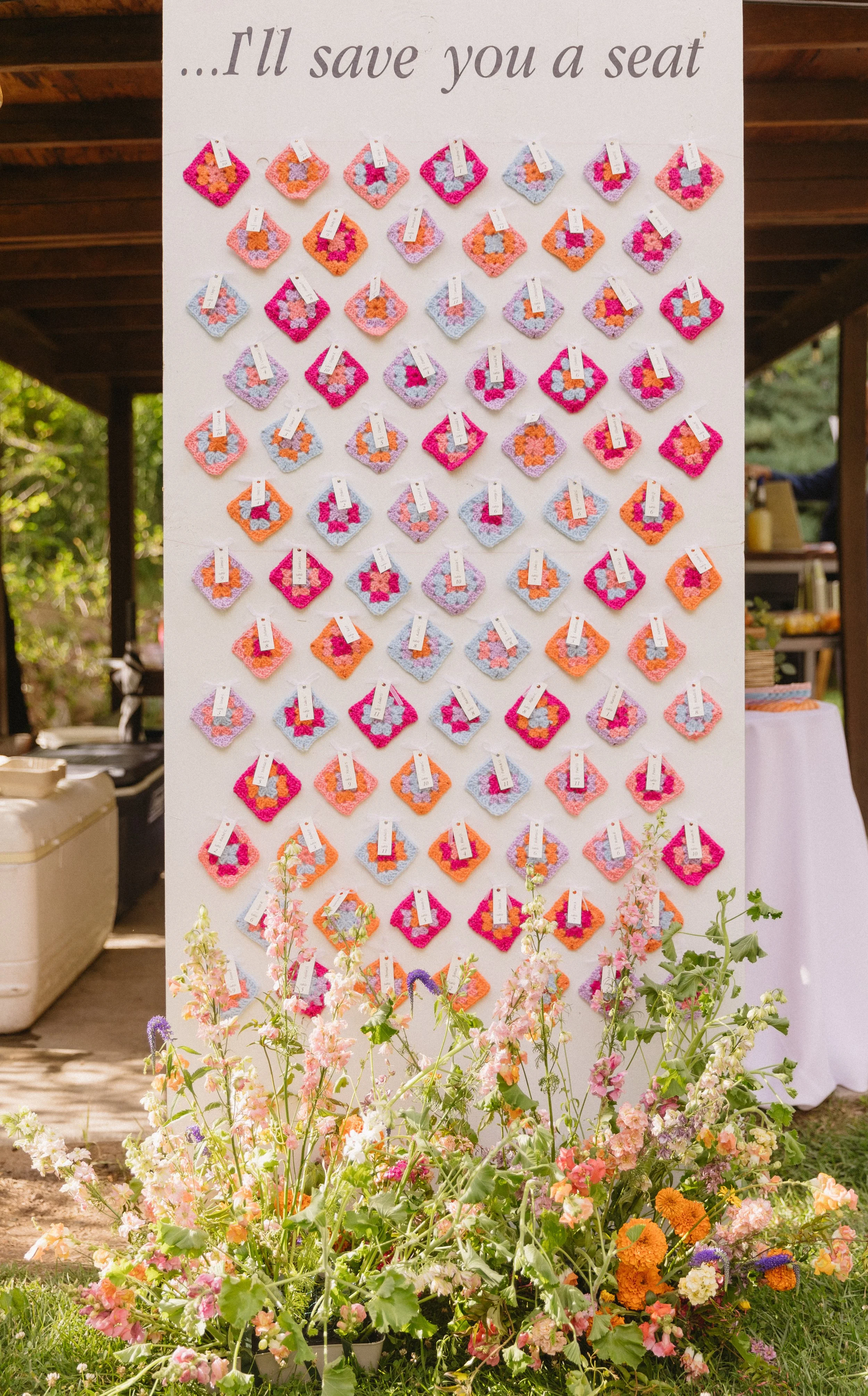 Colorful crochet squares with tags attached, arranged on a white display board with the phrase "...I'll save you a seat." in gray text at the top. In front of the display, there is a large, vibrant arrangement of assorted flowers.