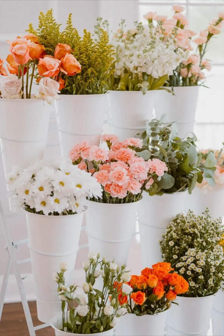 Arranged pink, white, and orange flowers in white buckets display on a white stand near a window.