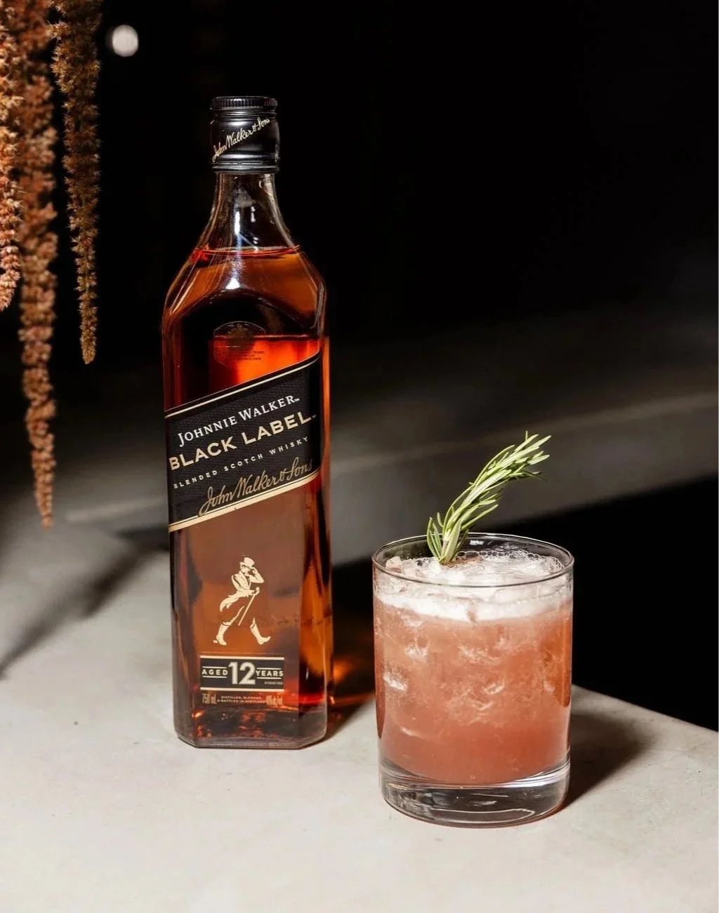 A bottle of Johnnie Walker Black Label blended Scotch whisky beside a glass of pink cocktail garnished with a sprig of rosemary, on a light-colored surface against a black background.