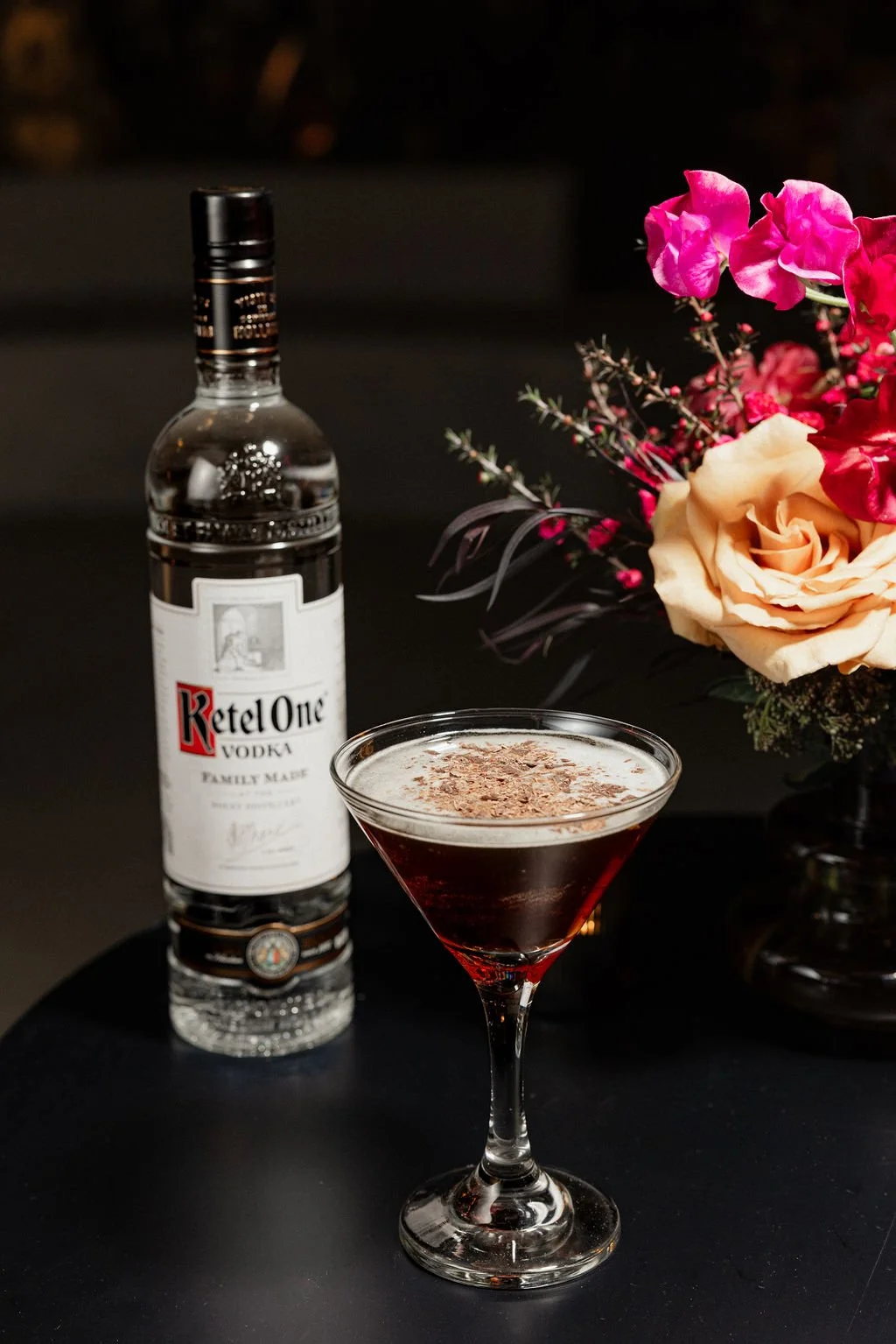 A cocktail in a martini glass garnished with chocolate shavings, a bottle of Ketel One vodka, and a bouquet of pink, peach, and red flowers on a black surface.
