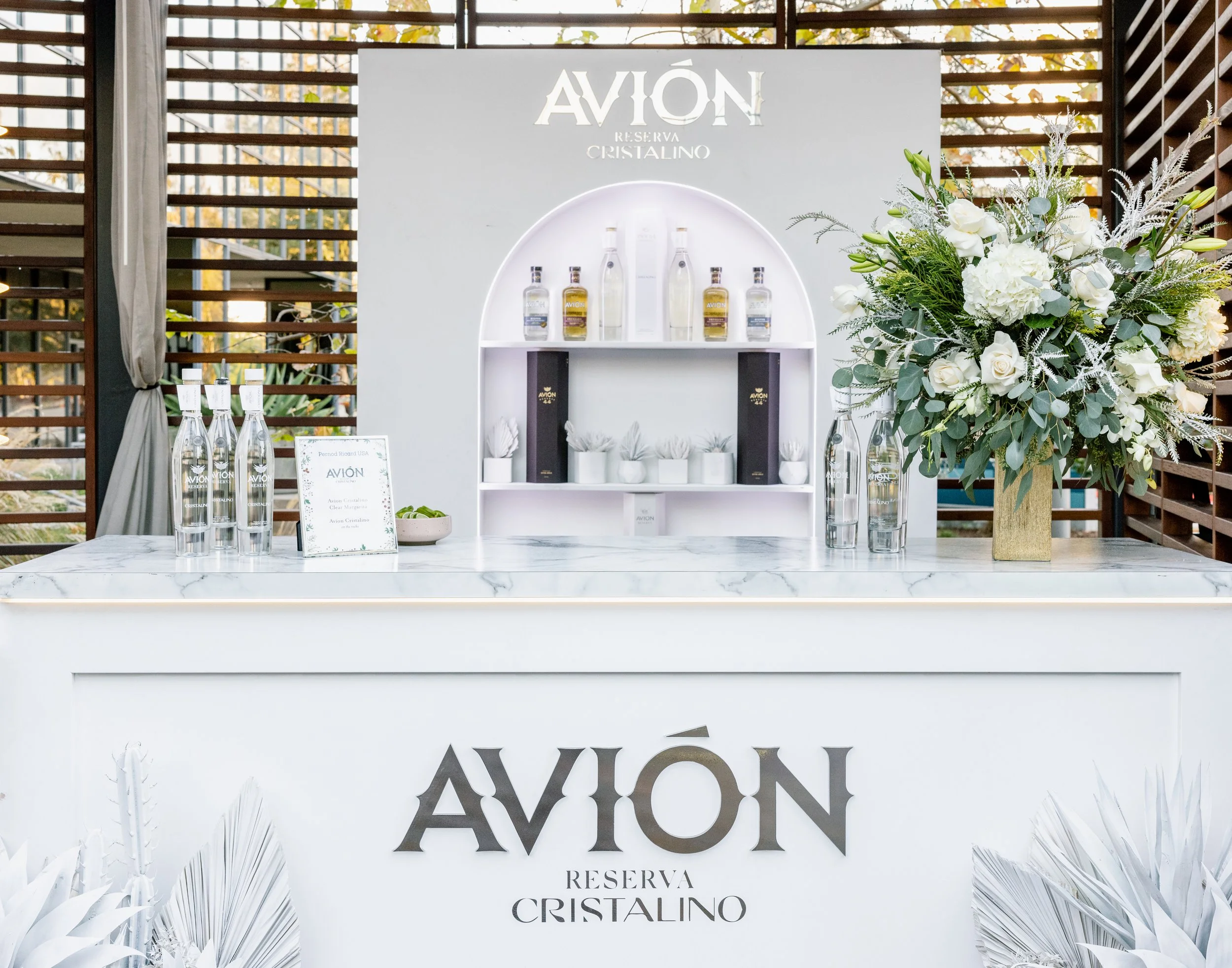 A display stand featuring bottles of Avión Reserva Cristalino tequila, a large floral arrangement with white flowers and greenery, and a sign on a marble countertop with the Avión logo and some text, set in an outdoor or semi-outdoor area with wooden slats and trees in the background.