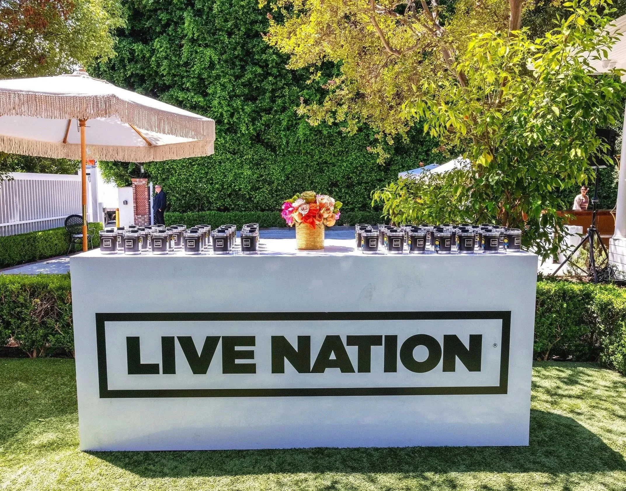 Outdoor scene with a white sign reading "LIVE NATION" in black letters, set in a grassy area with trees and shrubs, a table with black cans featuring the "LIVE NATION" logo, a large umbrella with fringe, and a person in the background.