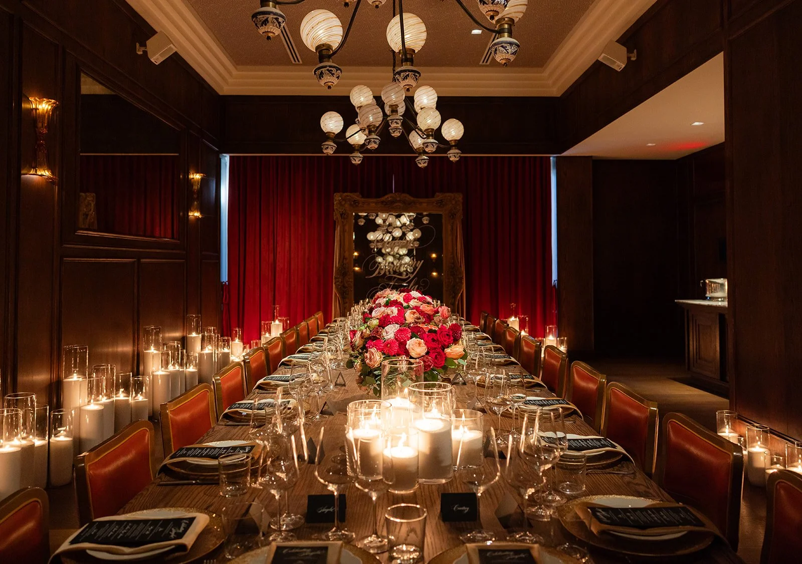Elegant dining room with a long wooden table decorated with a large floral centerpiece of red, pink, and peach flowers. The table is set with wine glasses, plates, and candles. The room has dark wood paneling, red curtains, and a chandelier with multiple globes hanging from the ceiling.
