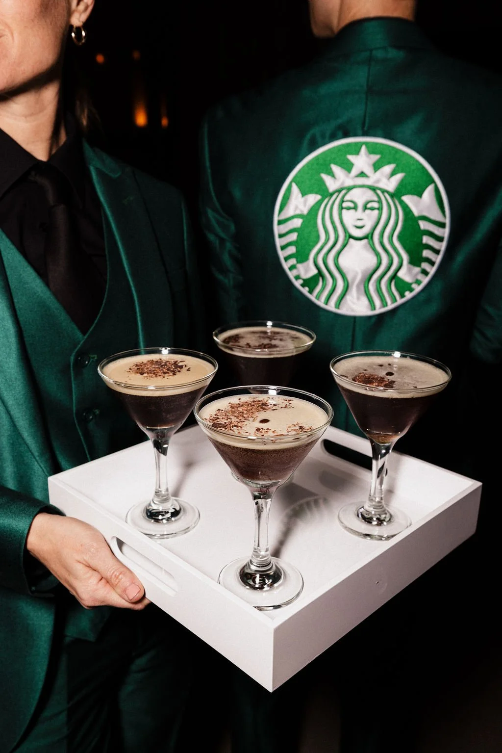 A person holding a tray of four espresso martini glasses with garnished drinks, standing next to another person wearing a green Starbucks jacket with the logo on the back.