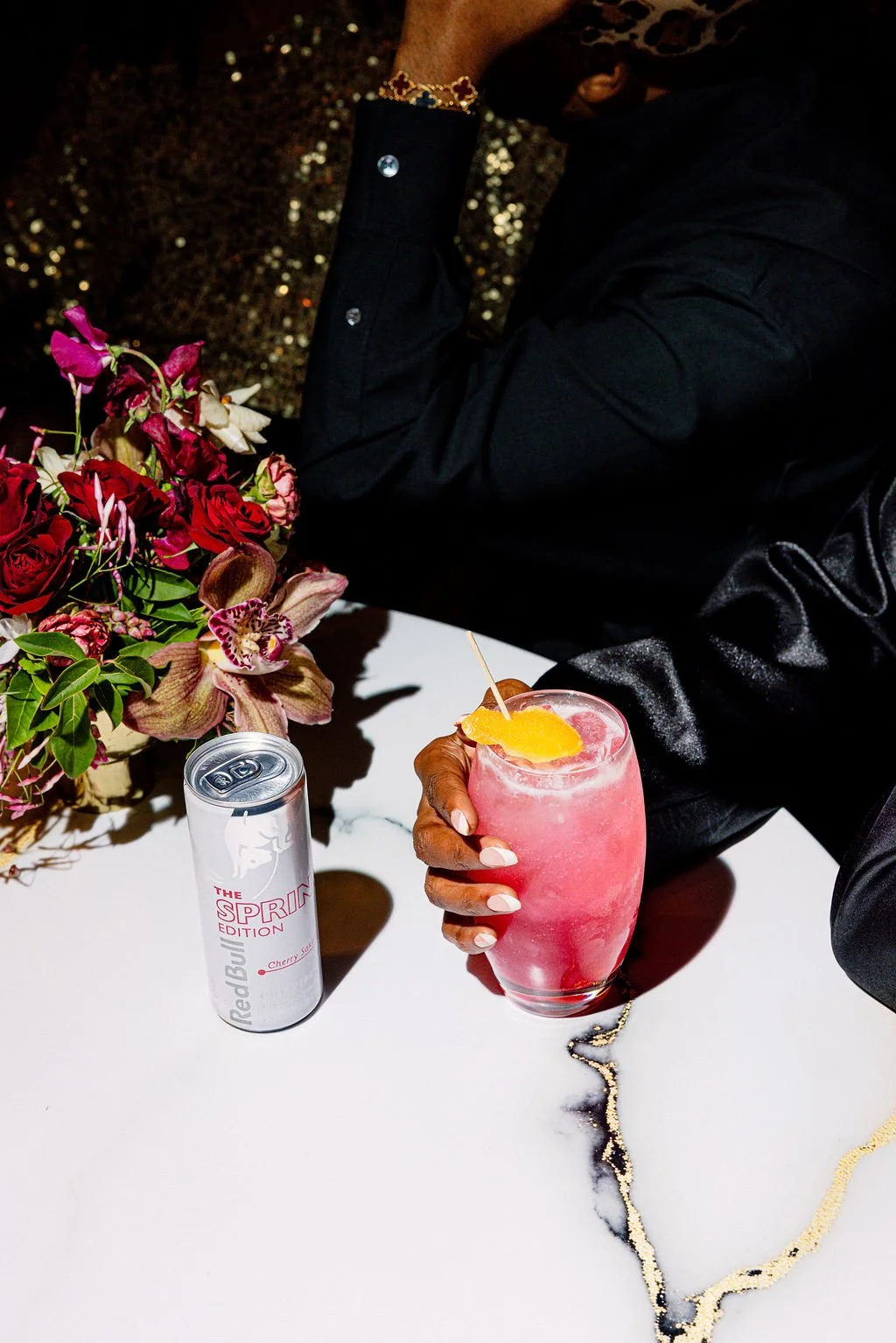 A person holding a pink cocktail with a slice of orange and a cherry, sitting at a table with a flower arrangement, a can of Red Bull energy drink, and a black purse, with a sparkly black surface in the background.