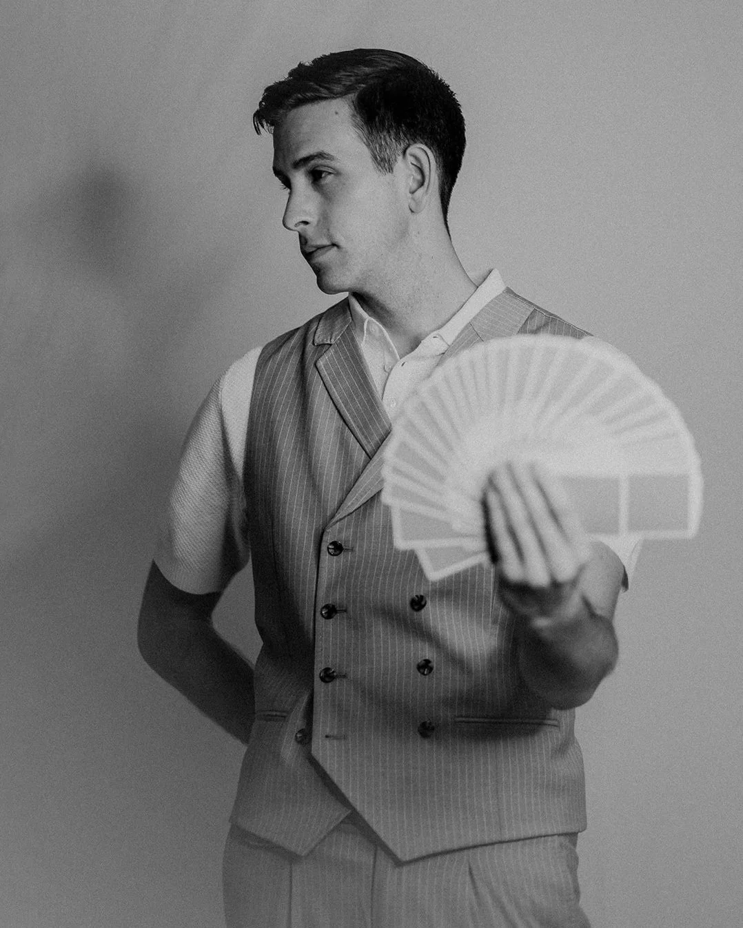 A man with dark hair in a suit holding playing cards in his right hand, looking to the left in a black-and-white photo.