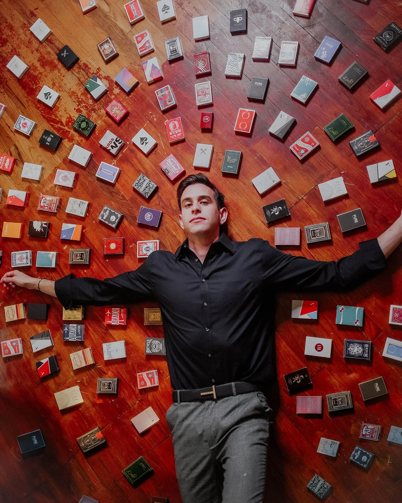 Young man with dark hair, wearing a black shirt and gray pants, lying on a hardwood floor surrounded by scattered cigarette packs.