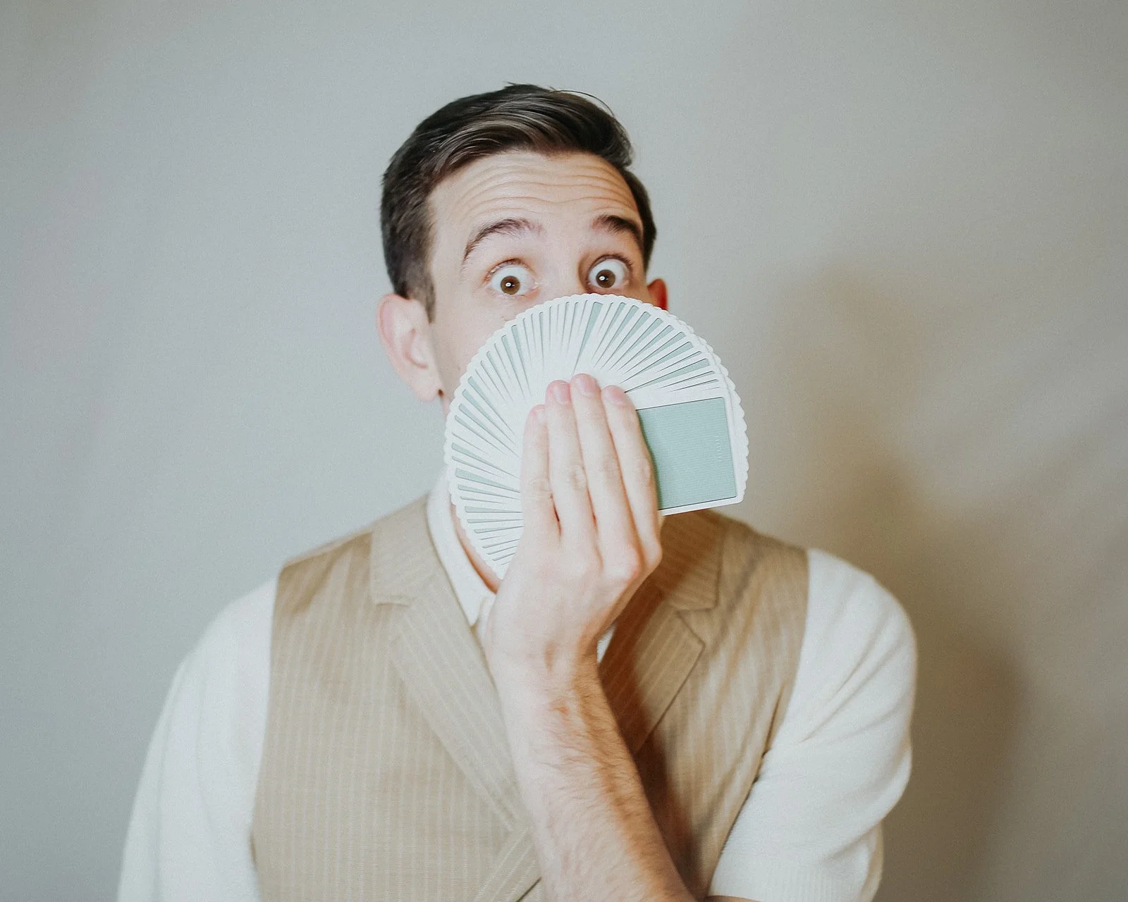 A man with short dark hair holds a fan of playing cards in front of his face, covering his nose and mouth, while looking at the camera with wide eyes.