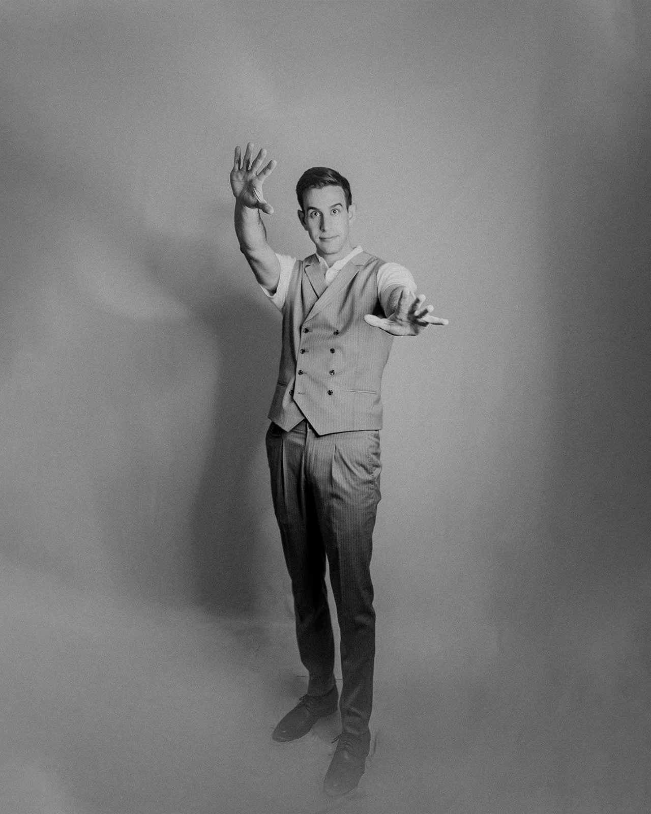 A black-and-white photograph of a young man in vintage attire, wearing a light-colored, double-breasted vest and matching trousers. He is standing against a plain background, with one arm raised and open hand near his head, and the other arm extended outward with an open hand. He has a neutral facial expression and neatly combed hair.