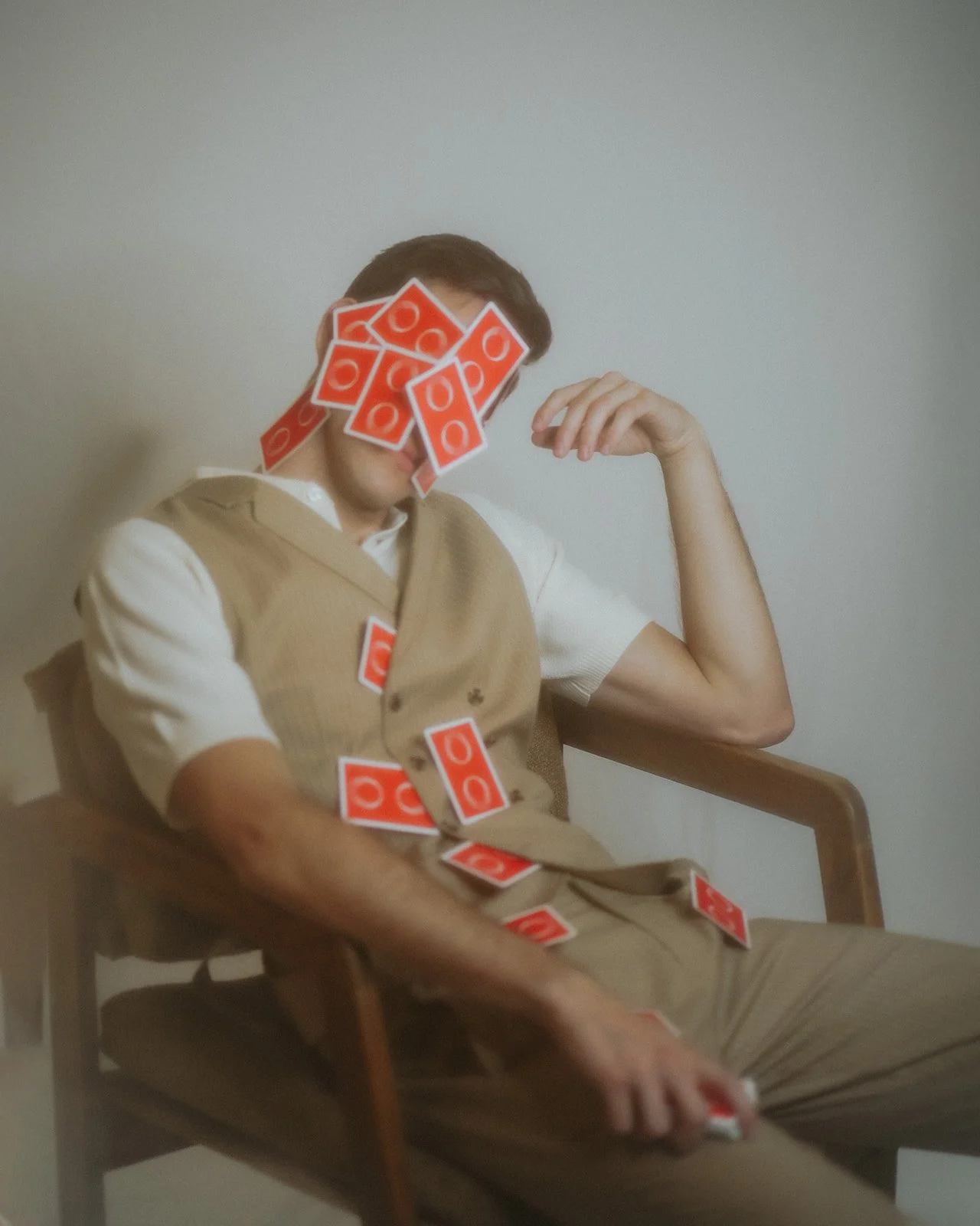 A person sitting on a wooden chair with their face covered by a pile of red and white playing cards. They are relaxed, with one arm resting on their knee and the other arm raised near their head. The individual is dressed in a beige vest over a white short-sleeved shirt.