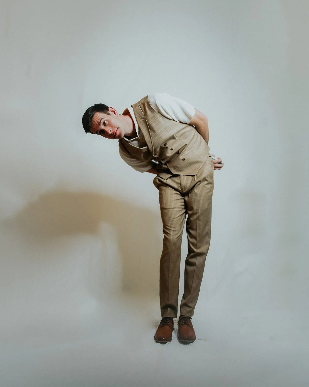 A man in beige pinstripe suit bending forward with his hands behind his back, looking at the camera against a plain light-colored background.