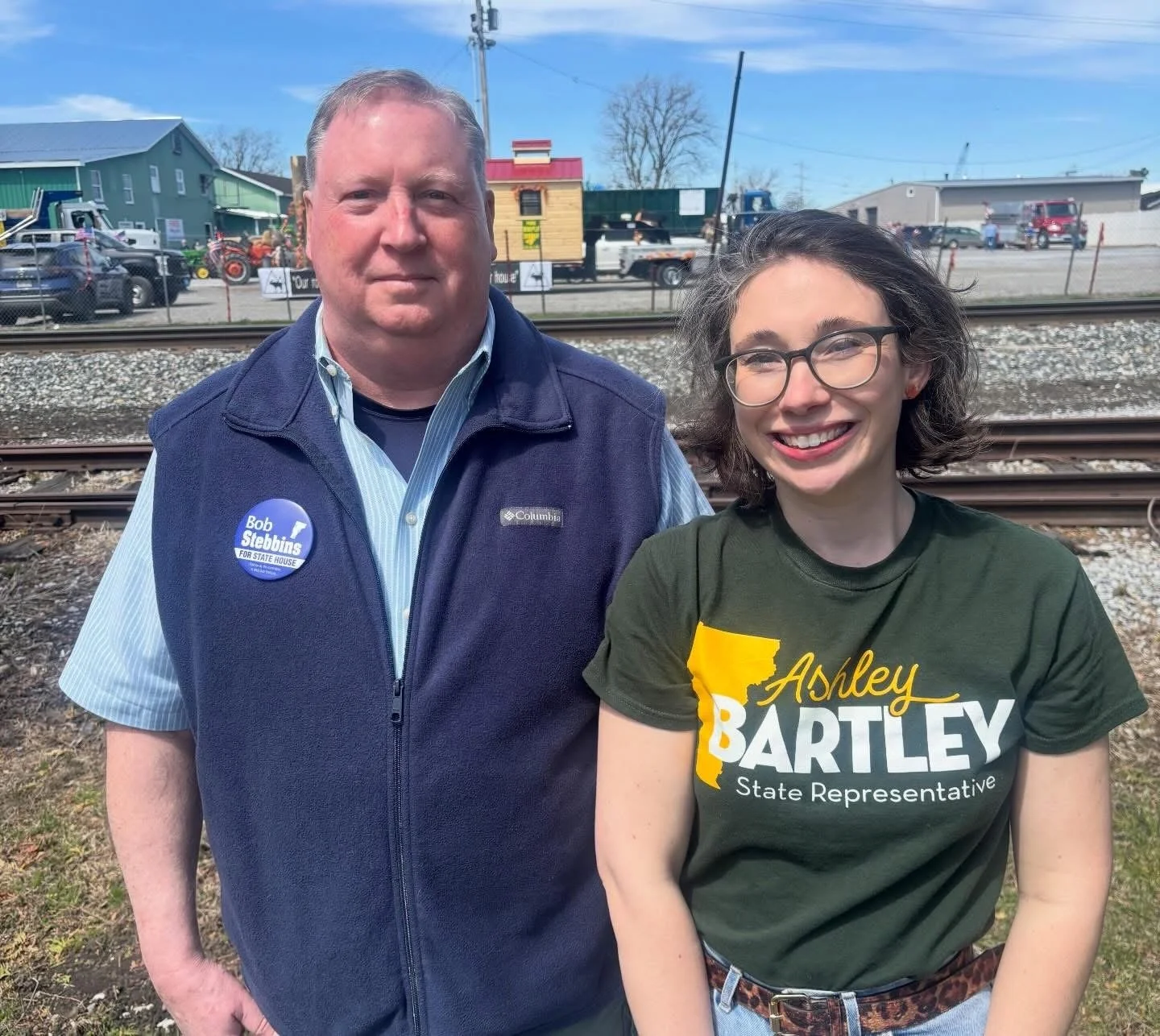 A man and a woman standing outdoors near train tracks; the man is wearing a blue vest with a badge that says Bob Stebbins for State House, and the woman is wearing a green shirt that says Ashley Bartley State Representative.