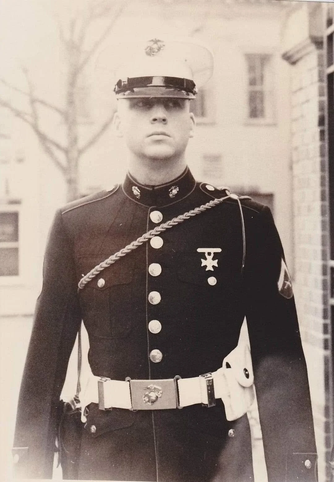A male U.S. Marine in dress uniform standing outdoors, wearing a cap, dark jacket with buttons, and a medal on his left chest, with a white belt and decorative cord over his shoulder.