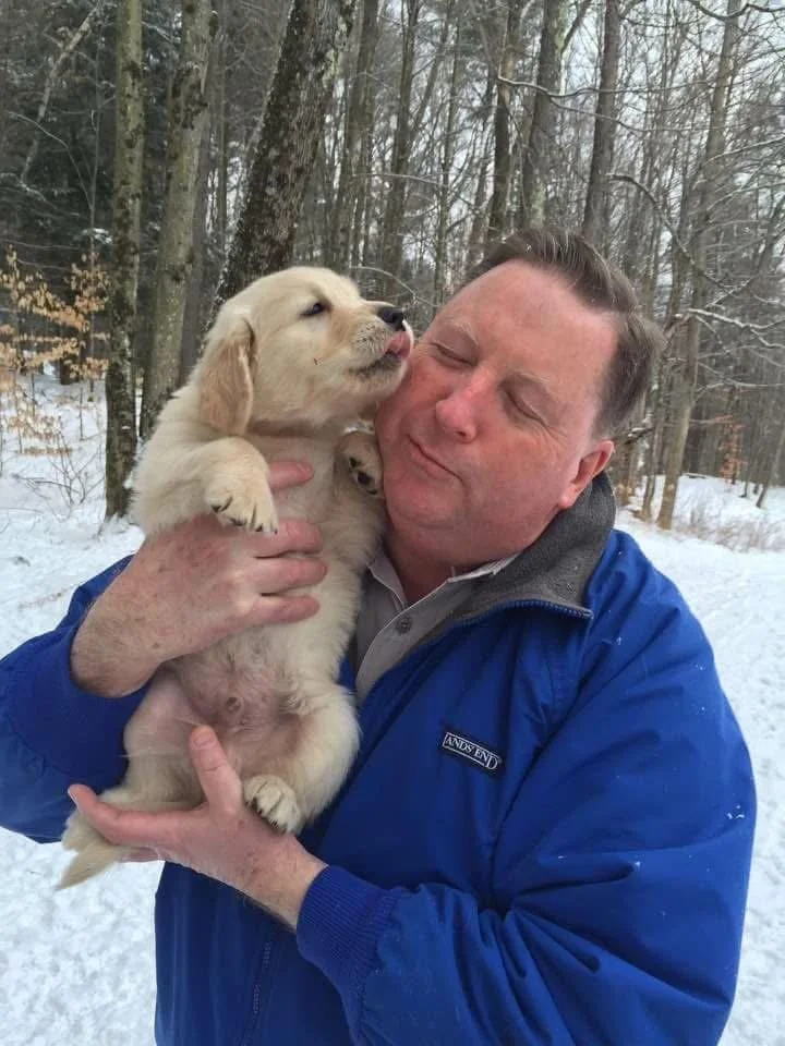A man in a blue jacket holds a golden retriever puppy that is licking his face in a snowy forest.