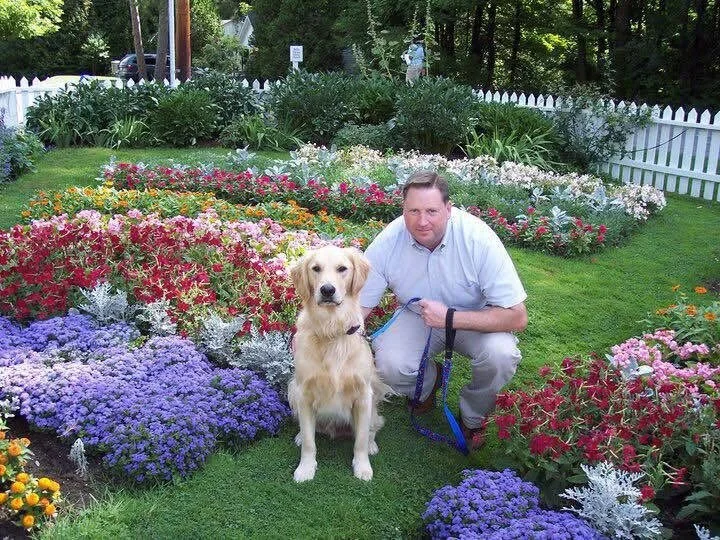 A man kneeling next to a golden retriever in a colorful flower garden with a white picket fence and trees in the background.
