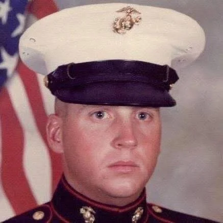 A young Marine in dress uniform with a white hat, black band, and Marine Corps emblem, standing in front of an American flag.