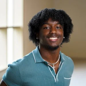 Close-up portrait of a smiling young man with curly black hair wearing a blue polo shirt and a necklace, standing indoors near a window.