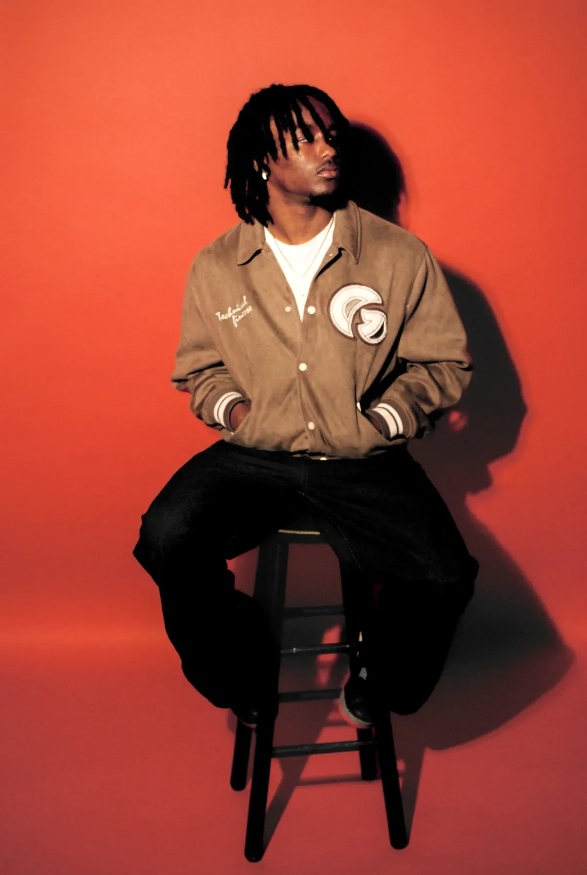 A man with dreadlocks sitting on a black stool against a red background, wearing a brown jacket and black pants, with his hands in his pockets, casting a shadow on the wall.