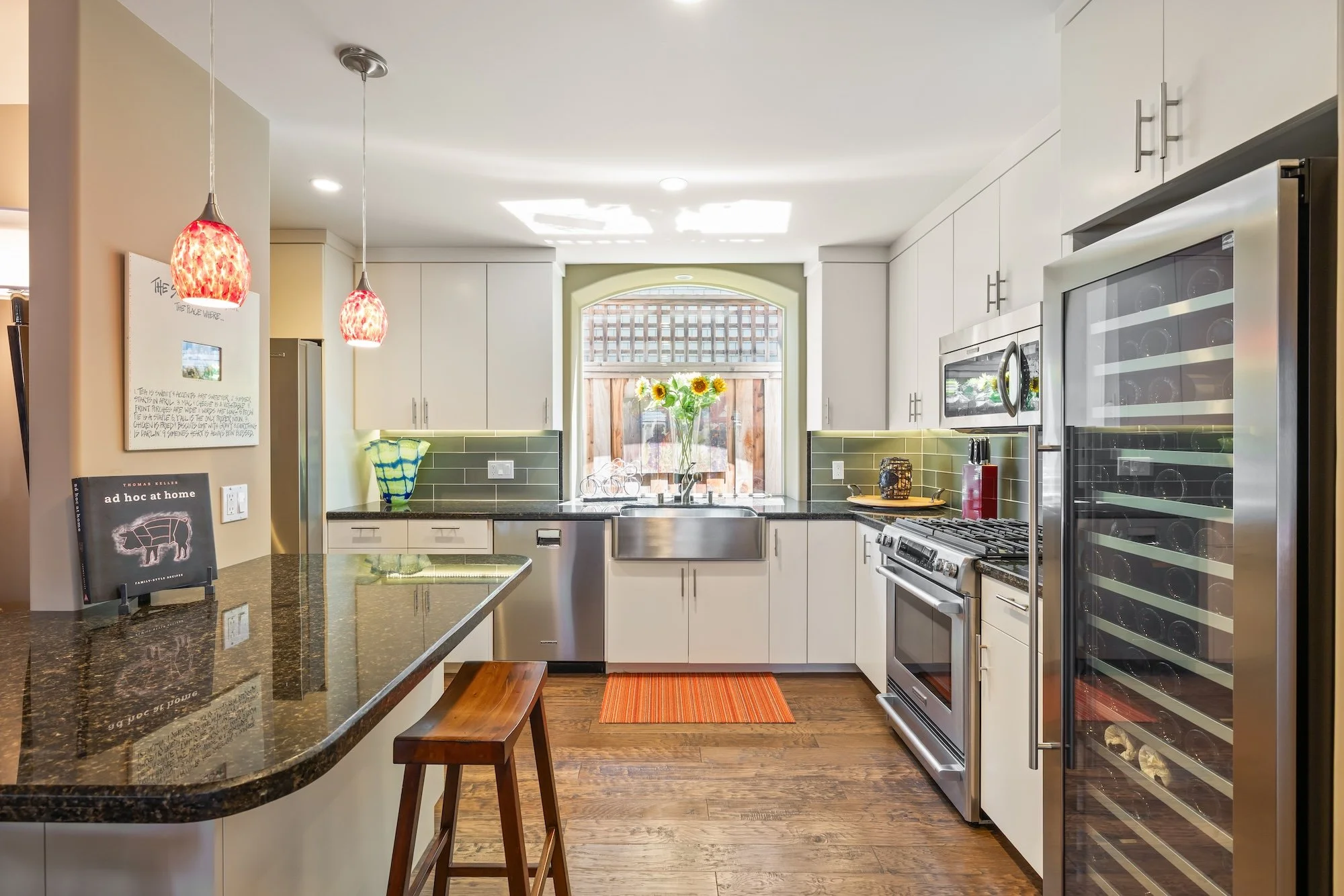 a modern kitchen with white cabinets, stainless steel appliances, a green tiled backsplash, a window with blinds, a vase of sunflowers on the windowsill, hanging pendant lights, a book on the counter, and wooden flooring.