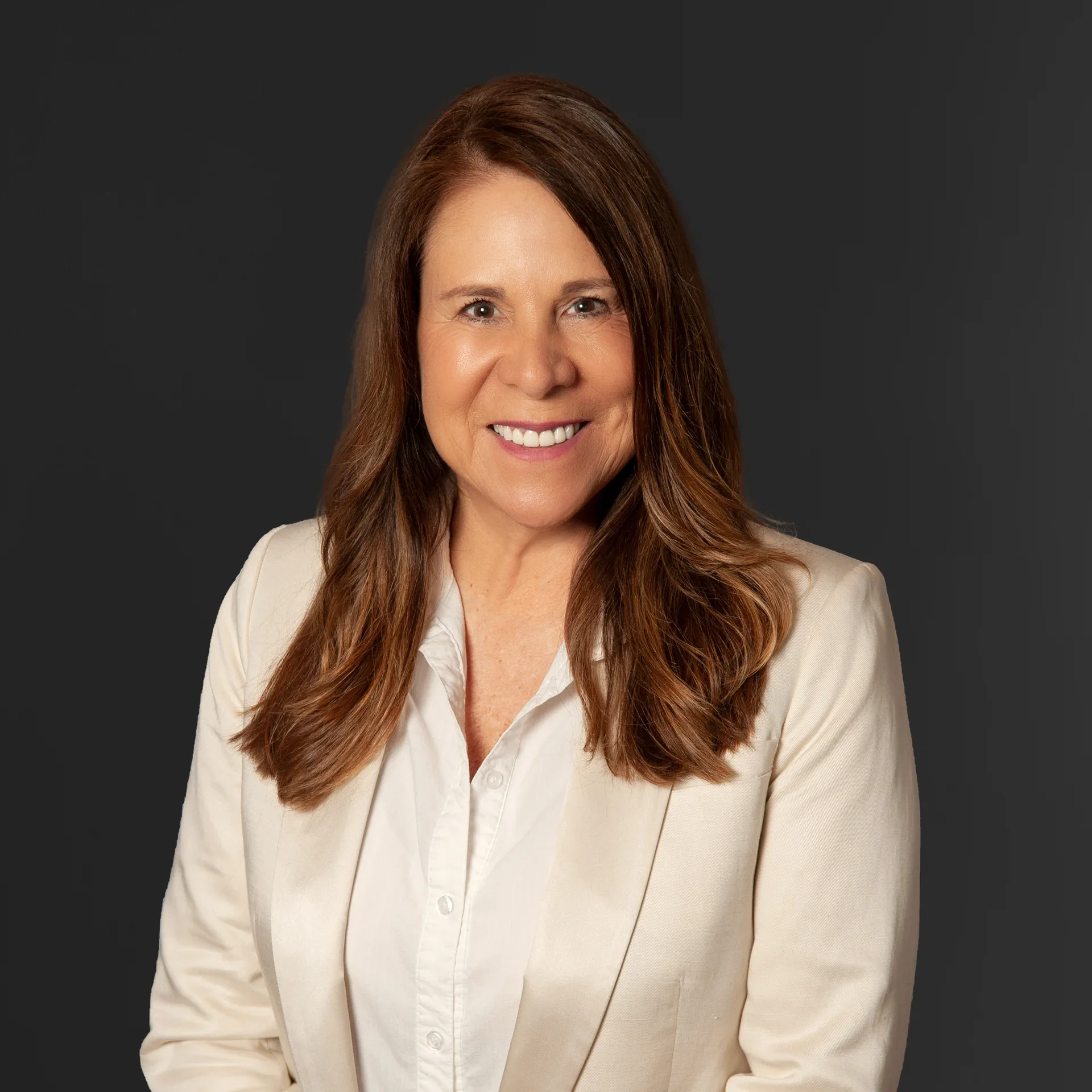 A woman with long reddish-brown hair, wearing a cream blazer and a white shirt, smiling against a dark gray background.