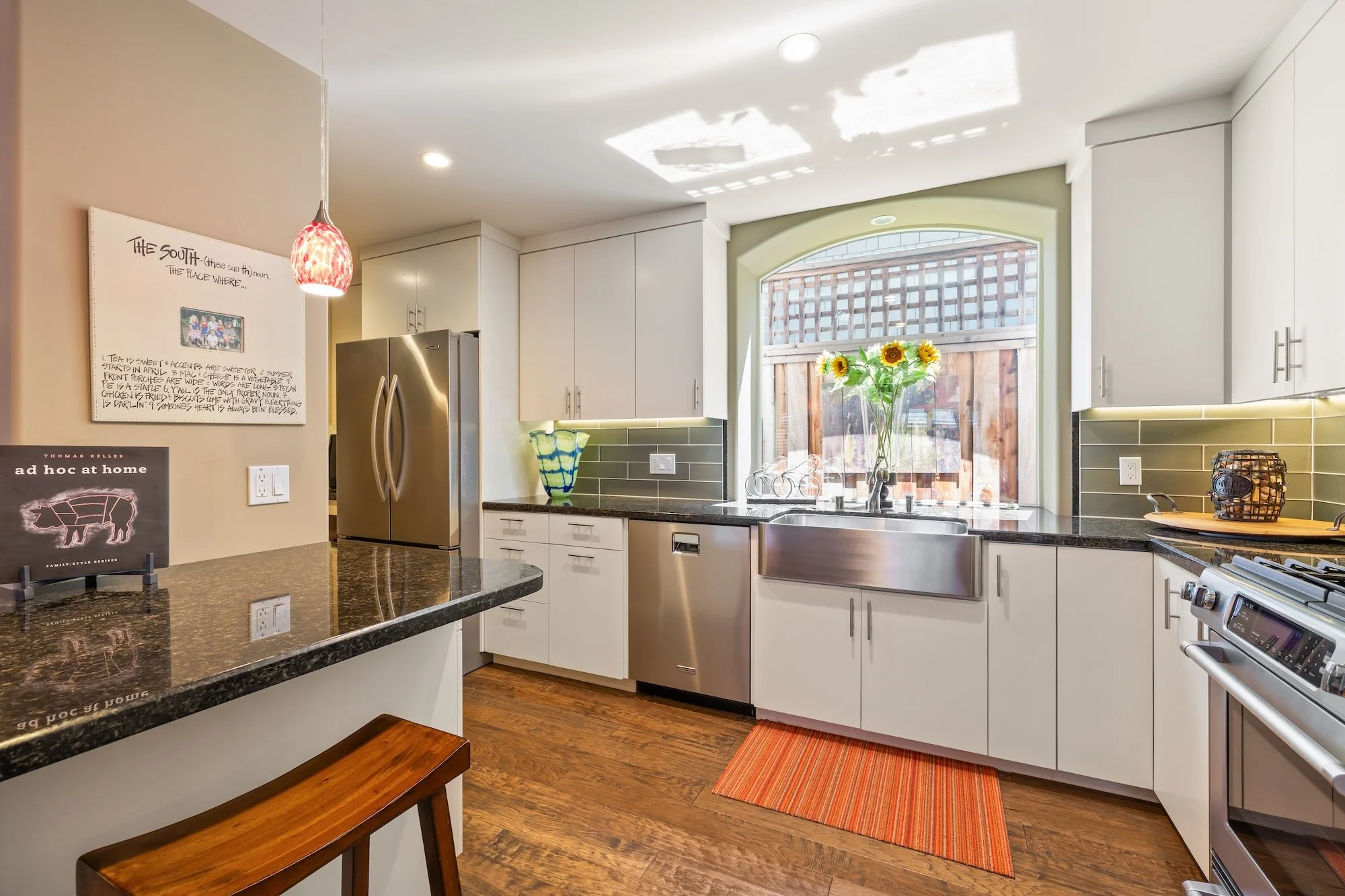 Modern kitchen with white cabinets, black granite countertops, stainless steel appliances, sunflower arrangement by a large window, and colorful decor.