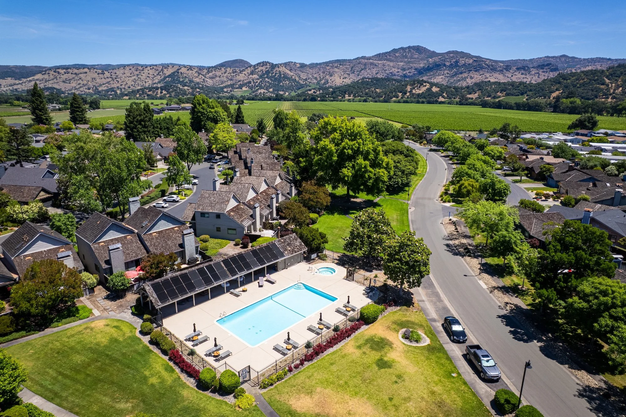 A residential neighborhood with houses, a swimming pool, and a clubhouse in the foreground, surrounded by trees and green lawns, with agricultural fields and mountains in the background under a clear blue sky.