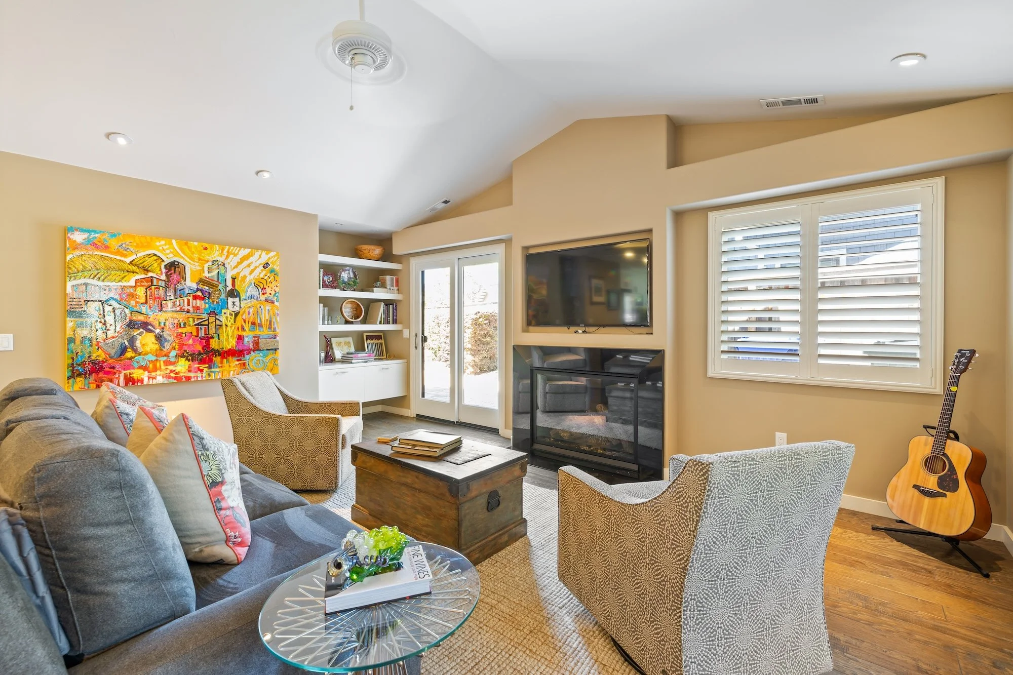 Living room with beige walls, a gray sofa with colorful pillows, two patterned armchairs, a wooden coffee table, a wall-mounted TV above a fireplace, large windows with white shutters, bookshelf with décor, and an acoustic guitar in the corner.