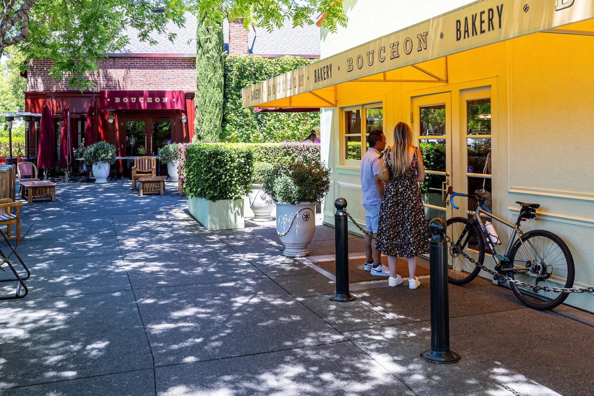 Two people are standing at the entrance of a bakery named Bouchon. The bakery has a yellow exterior with a beige awning. There are bicycles parked nearby, and outdoor seating with tables and umbrellas. Trees and greenery are visible, providing shade.