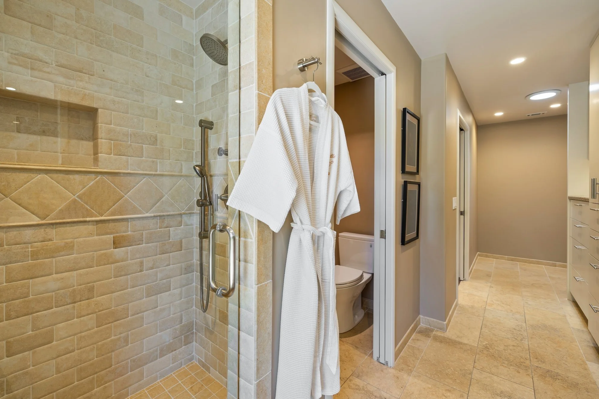 Bathroom with a walk-in shower with beige tiles, a robe hanging on a wall hook near the shower, and a toilet inside a small enclosed space with a door, with a hallway featuring beige walls and framed pictures, ceiling lights, and a marble floor.