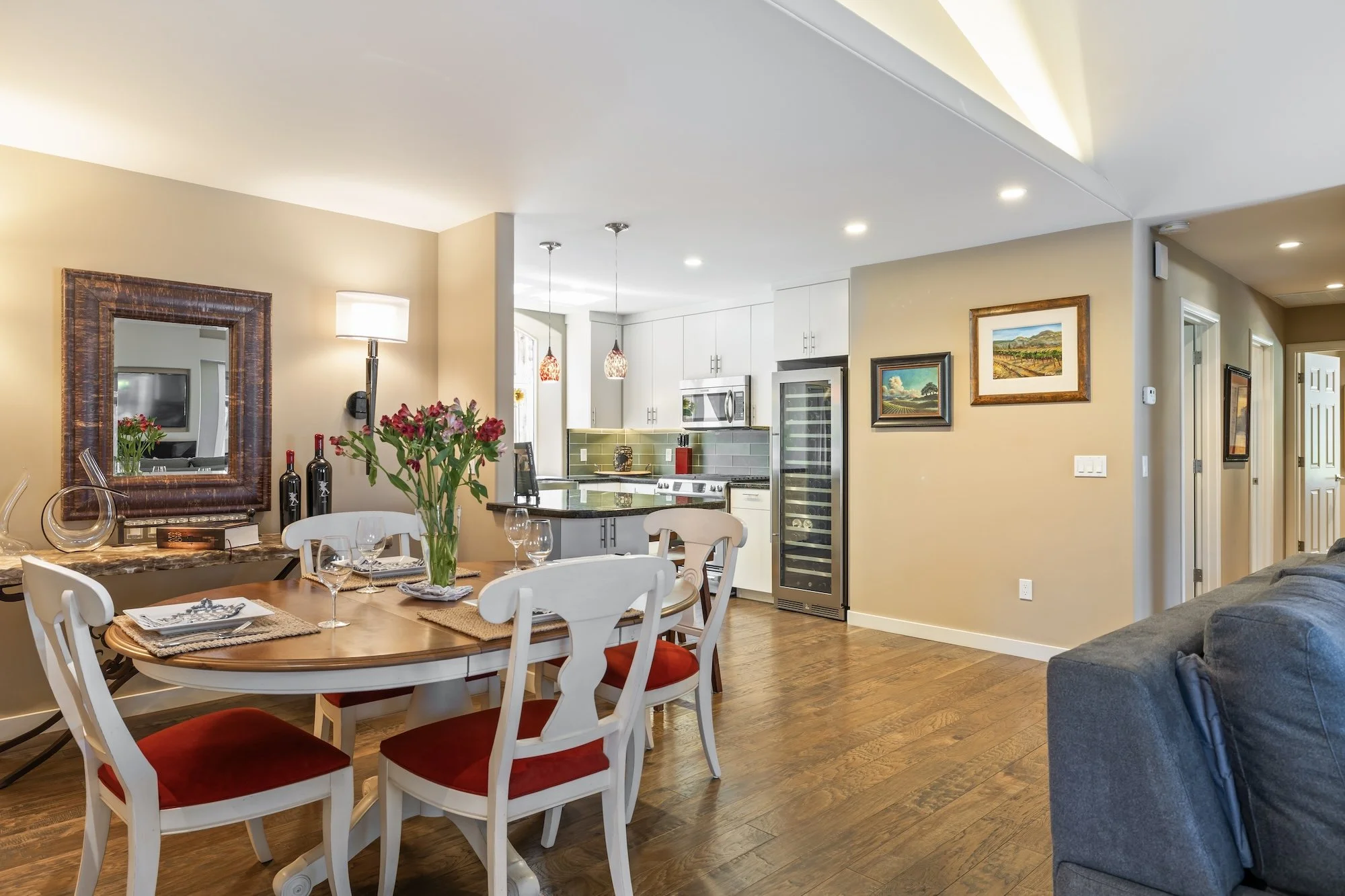 Open-concept living and dining area with a kitchen in the background. The dining table has a vase with pink and green flowers, and chairs with red cushions. The wall features framed paintings, and there is a mirror and decorative items on a side tabl