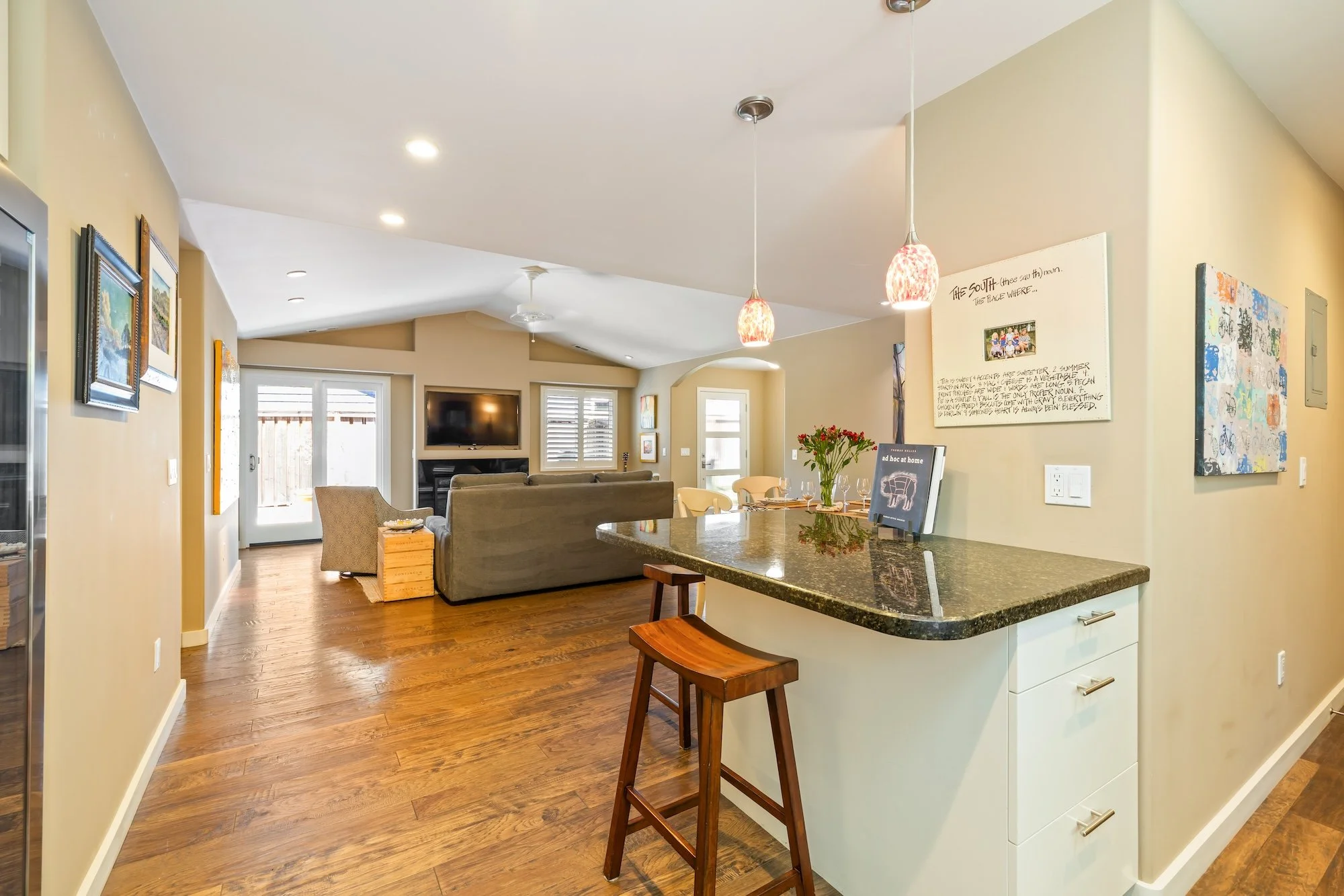 Open-concept living space with a kitchen island, granite countertops, and a cozy seating area with a TV. The room has hardwood floors, beige walls, and artwork, pendant lighting, and large windows allowing natural light.