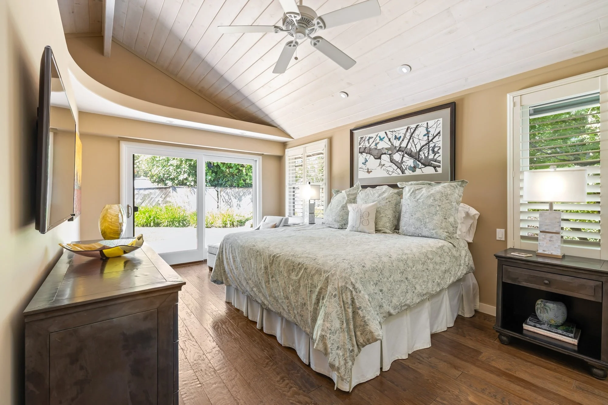A cozy bedroom with a vaulted white wood ceiling and ceiling fan. There's a large window with shutters showing greenery outside. The bed has light-colored, patterned bedding and multiple pillows. On each side of the bed, there are nightstands with la