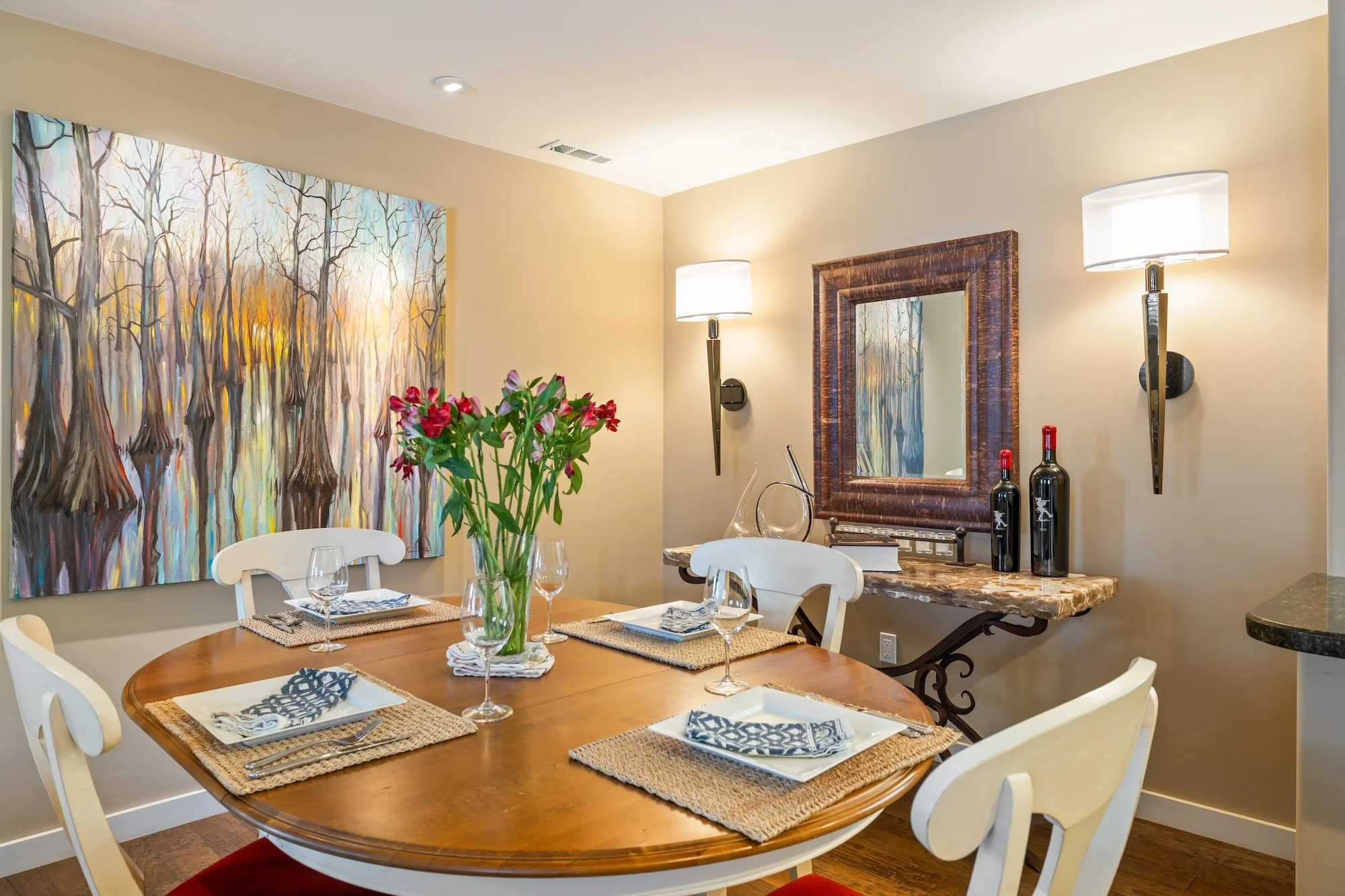 A dining room with a wooden table set for four with wine glasses, plates, and napkins, surrounded by white chairs. A large colorful painting of a forest scene dominates one wall, and a pair of tall modern lamps and a framed mirror are on another wall