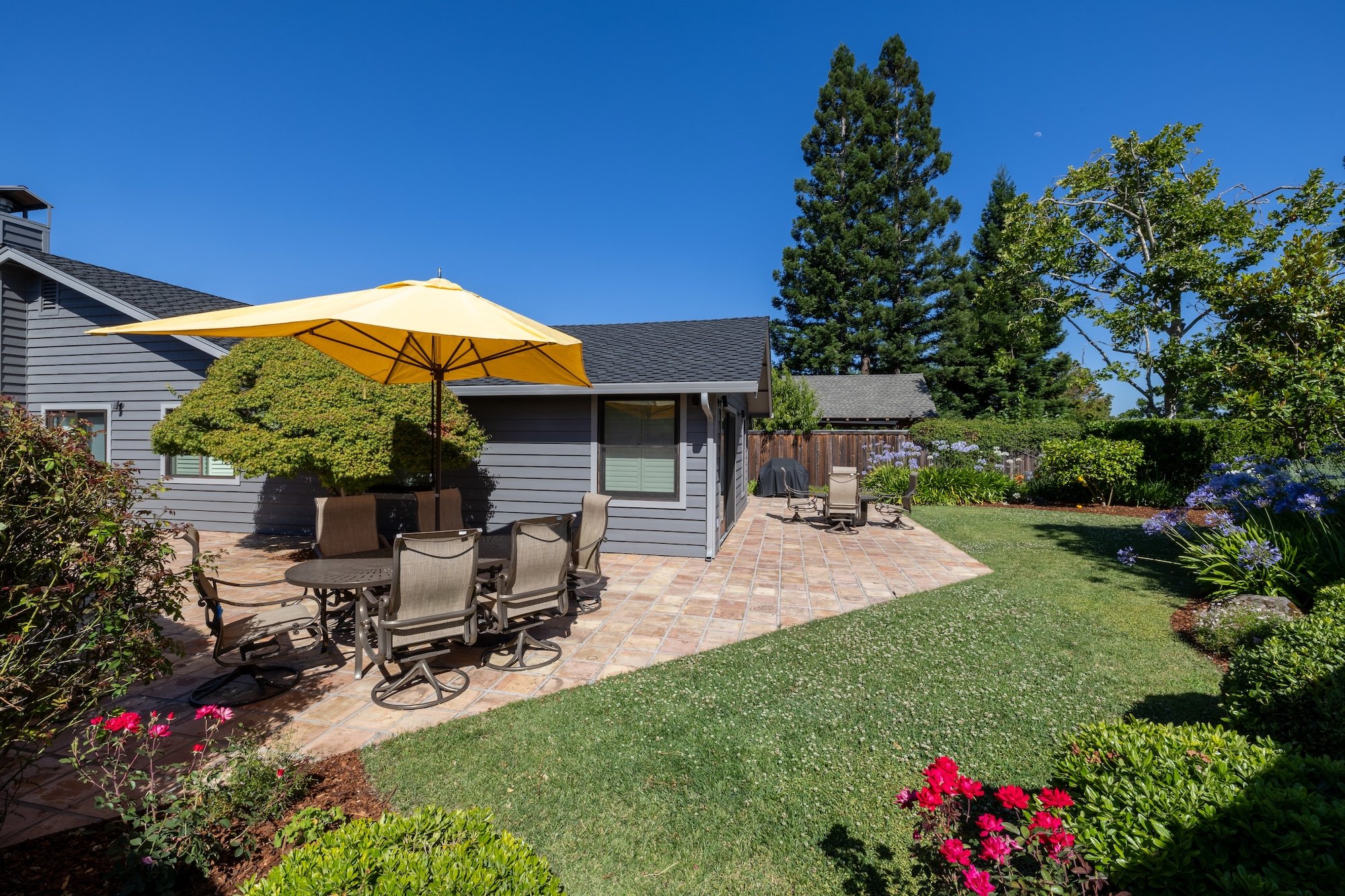 A backyard with brick patio and outdoor furniture, yellow patio umbrella, lush garden with colorful flowers and green trees, house with gray siding and a window, tall pine tree in the background, clear blue sky with a faint moon.