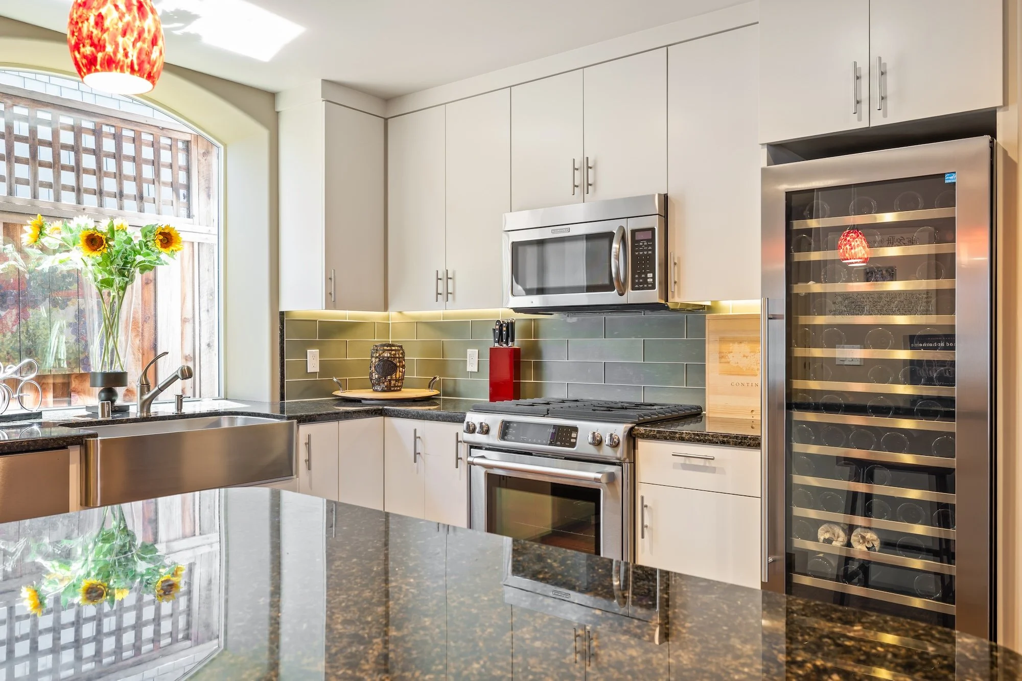 Modern kitchen with white cabinets, black granite countertops, a stainless steel stove, microwave, and wine cooler. A window with sunlight, a vase of sunflowers, and a red pendant light add color.