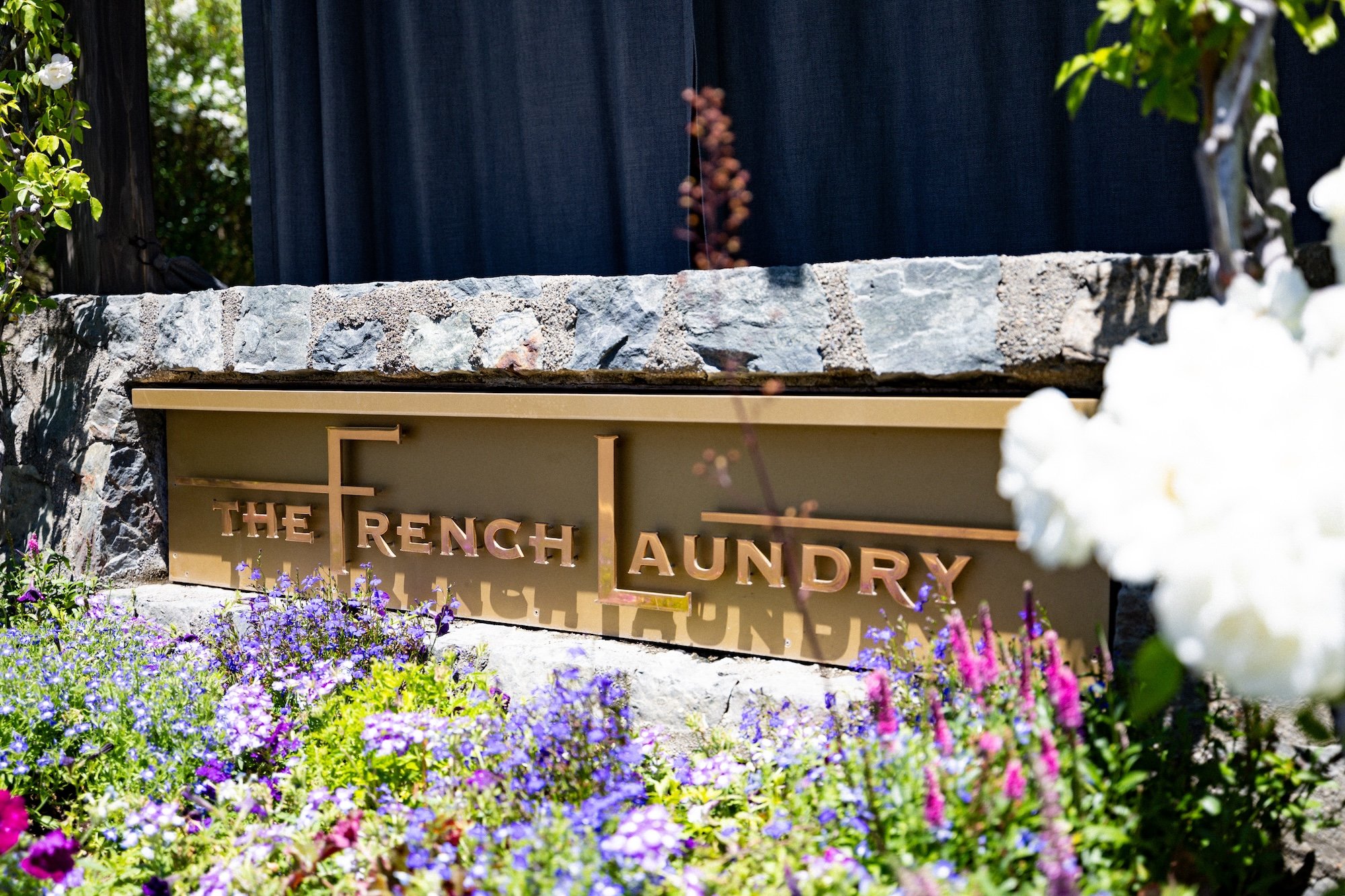 Sign for The Rench Aluminum Laundry in front of a stone wall, surrounded by purple and pink flowers, with trees and a dark curtain or fence in the background.