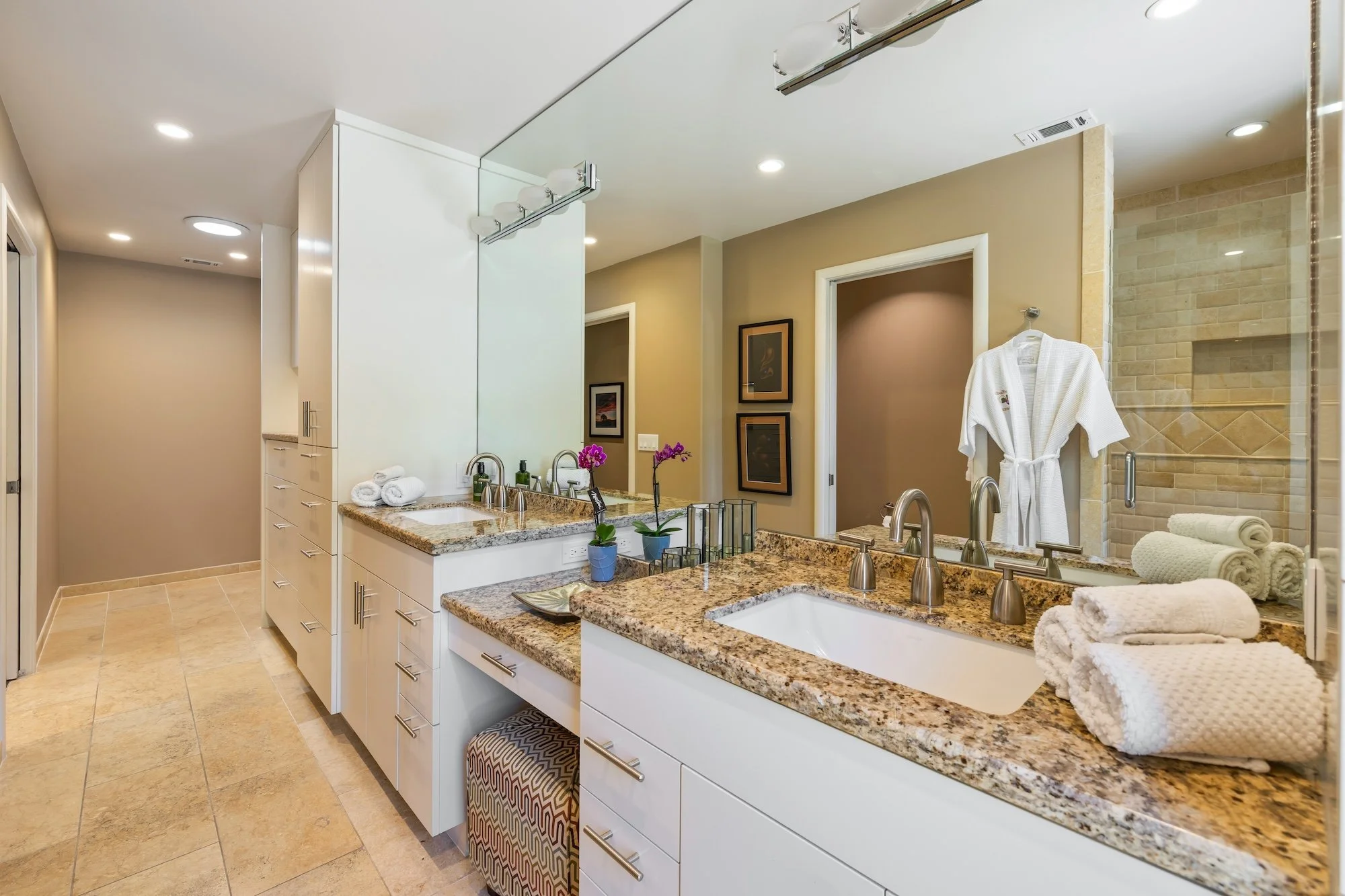 Bathroom with granite countertops, white cabinets, tiled floors, and a large mirror. Towels and robes are neatly arranged, with a shower area visible in the background.