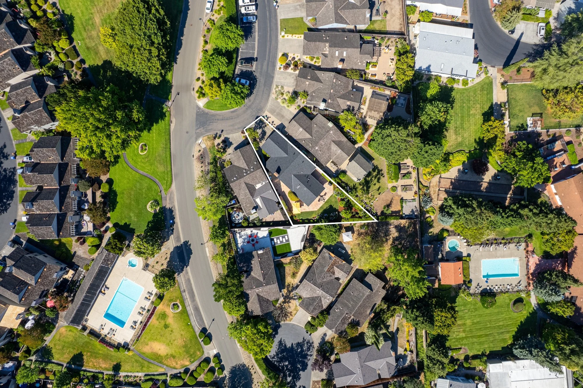 Aerial view of a suburban neighborhood with houses, trees, green lawns, and swimming pools.