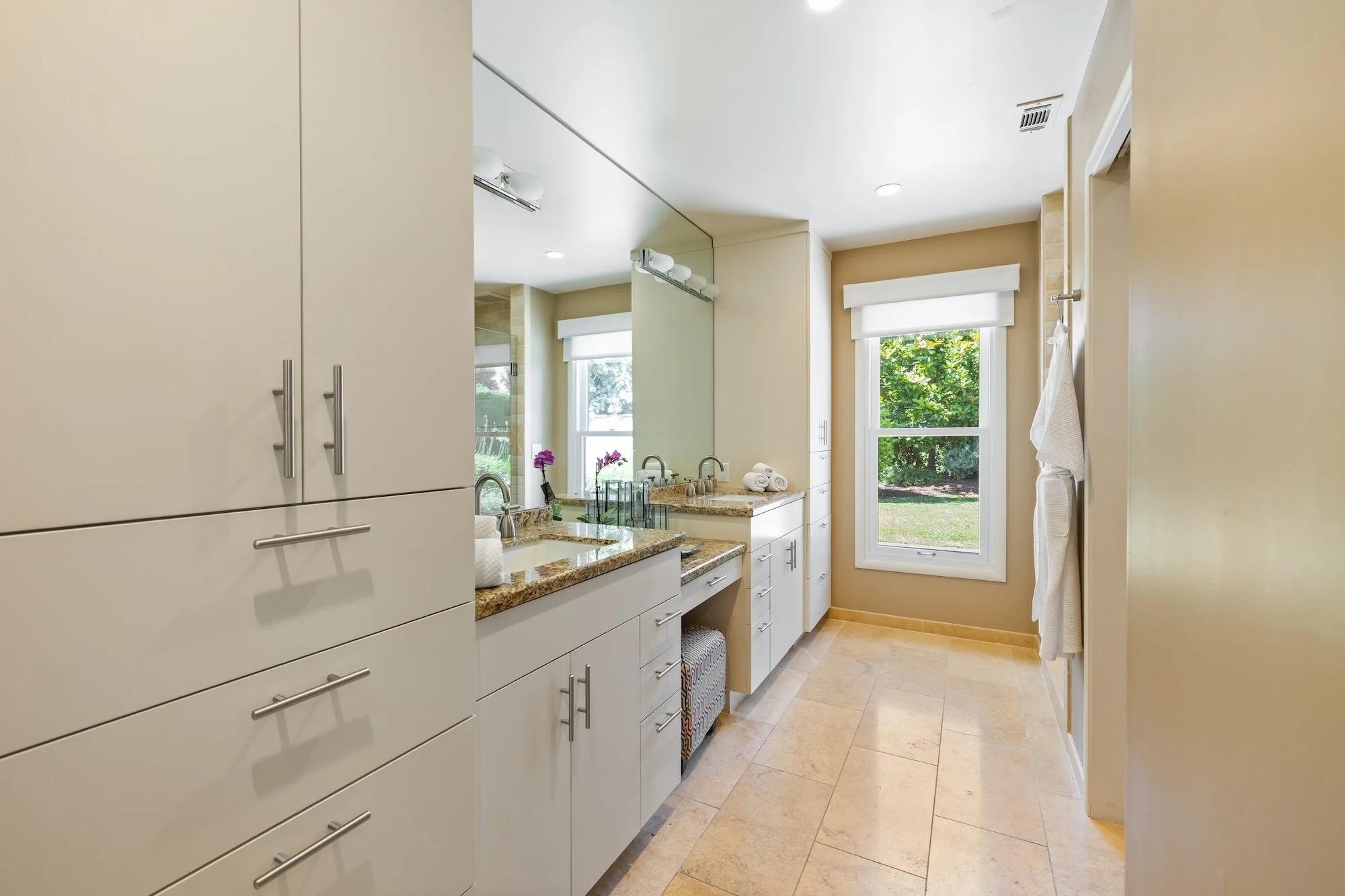 Bright bathroom with large window overlooking green trees, featuring white cabinets, granite countertops, and a mirror.