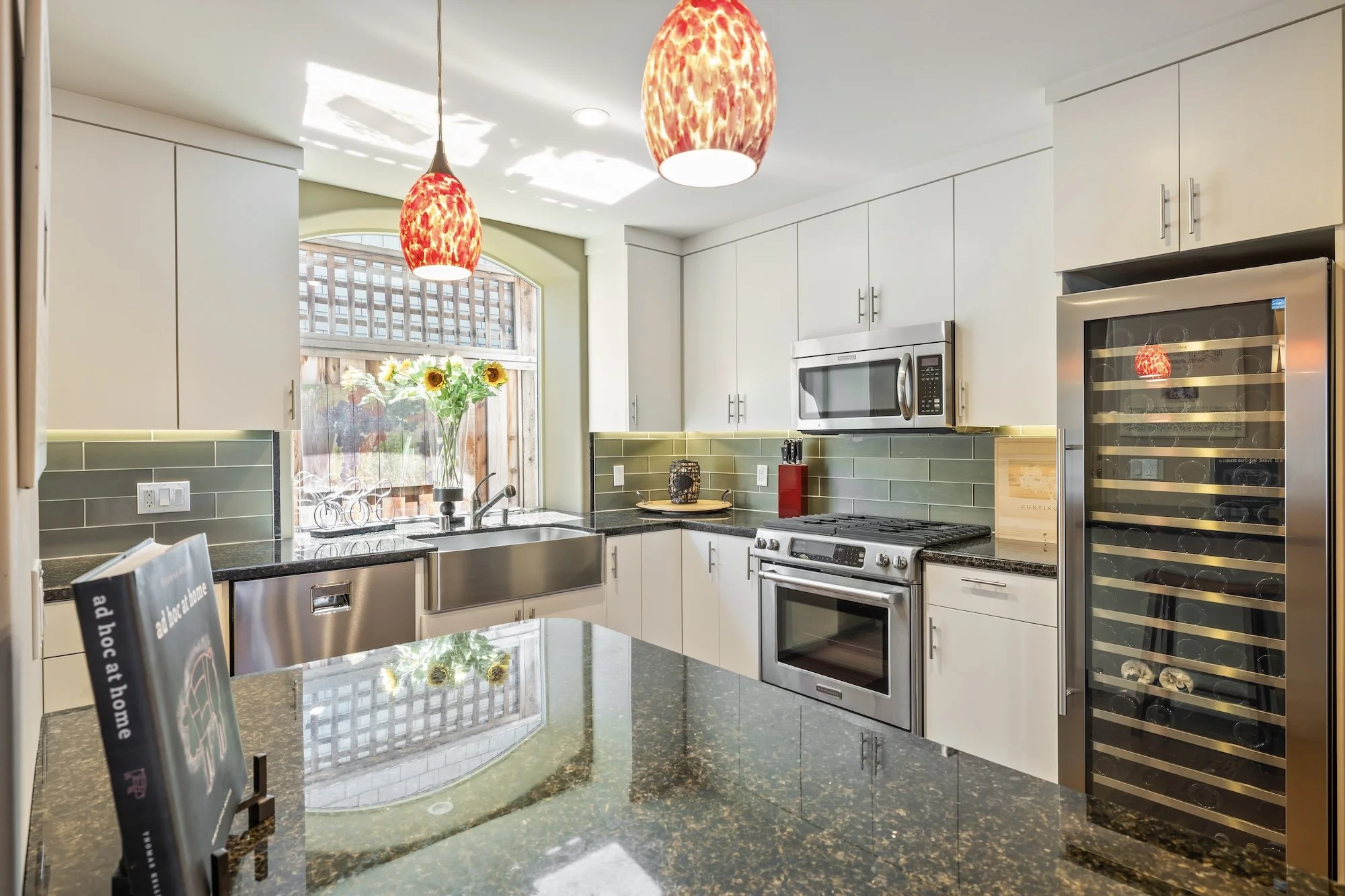 Modern kitchen with white cabinets, a stainless steel oven and microwave, a wine fridge, green tiled backsplash, a large window with sunlight, and two red pendant lights hanging from the ceiling.